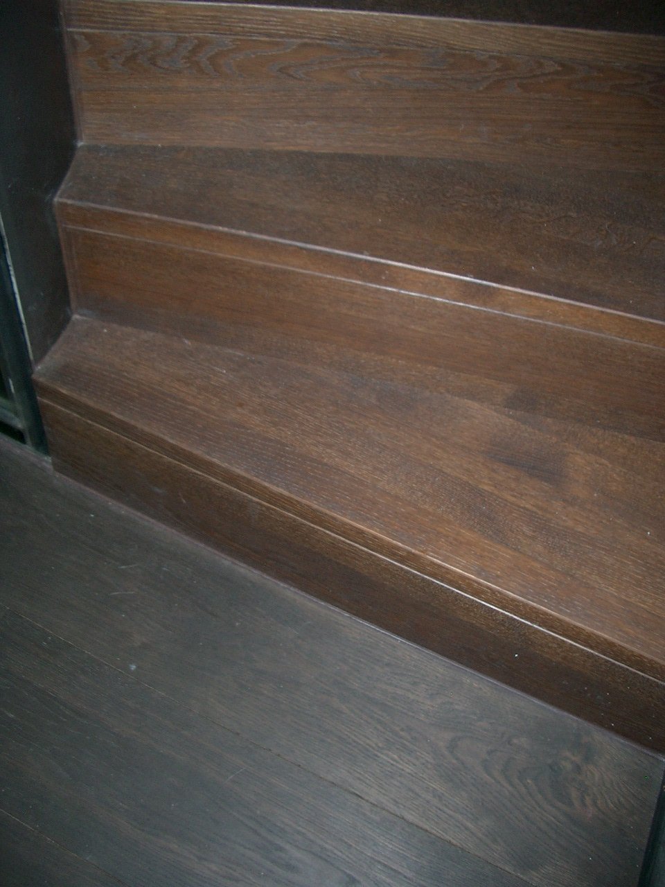 Close-up of wooden stairs with dark brown finish, showing the edge of the steps and the floor at the bottom.