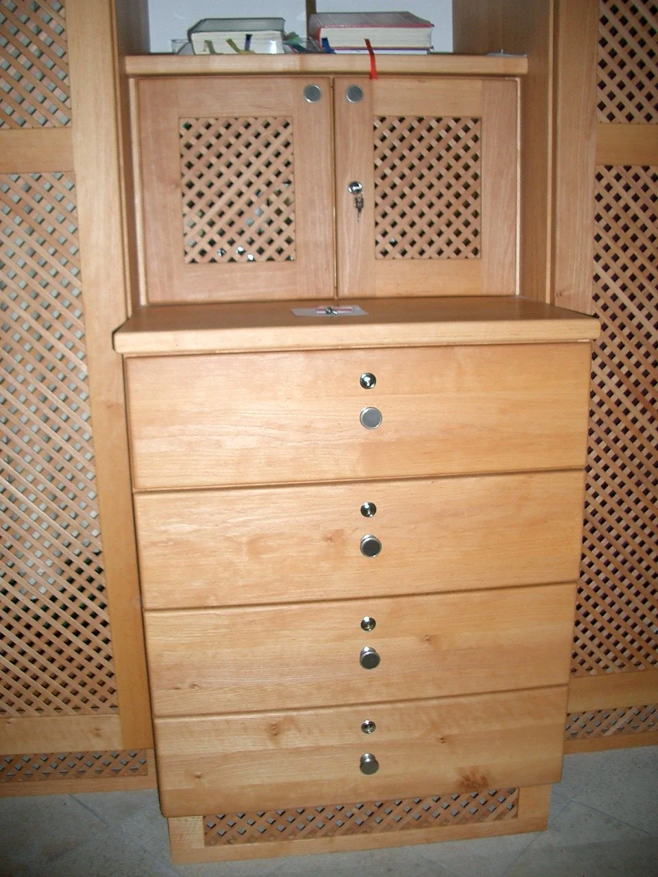 Wooden dresser with six drawers and a top cabinet with two doors, adorned with a keyhole and a couple of small round metal accents, placed in front of a wooden lattice partition, with books and a small container on top.