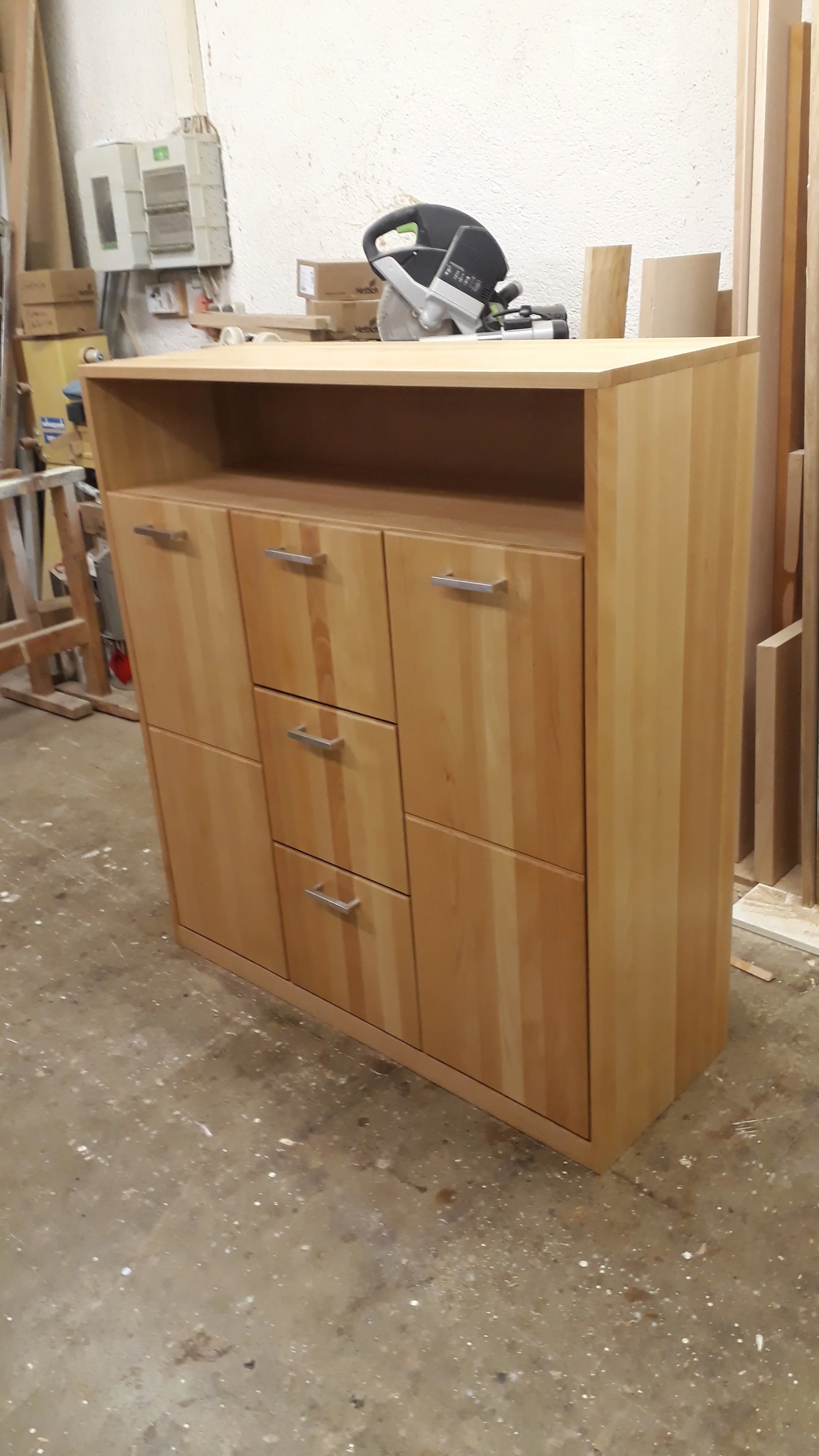 A wooden cabinet with multiple drawers and an open shelf, placed in a woodworking workshop with tools and materials around.