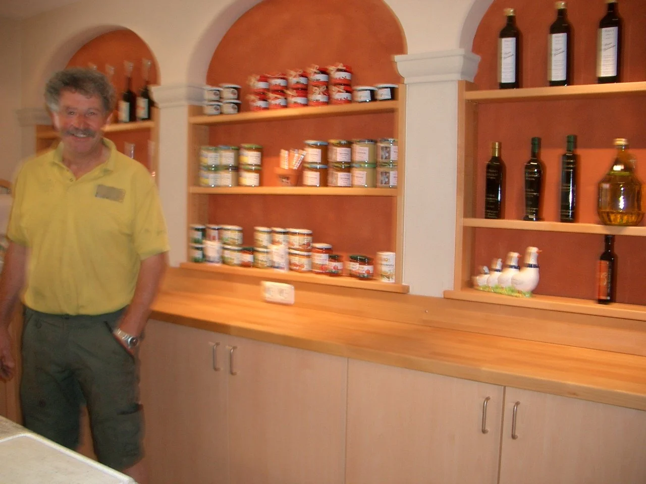 A man in a yellow shirt standing inside a store or kitchen with wood shelves holding jars and bottles.