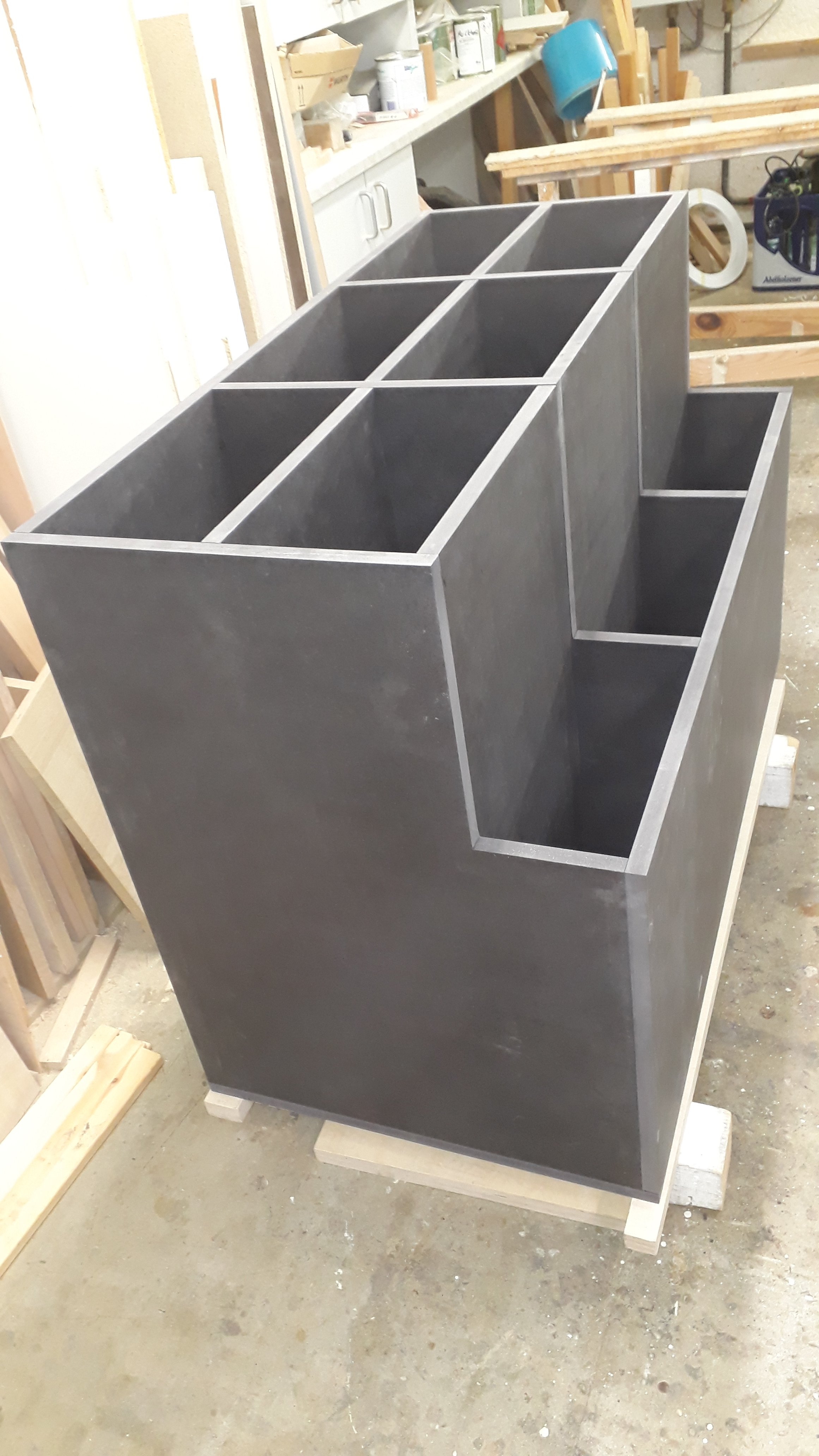 A black wooden dresser with multiple open compartments is in a woodworking shop, resting on small wooden blocks. The background shows shelves with various tools, supplies, and equipment.