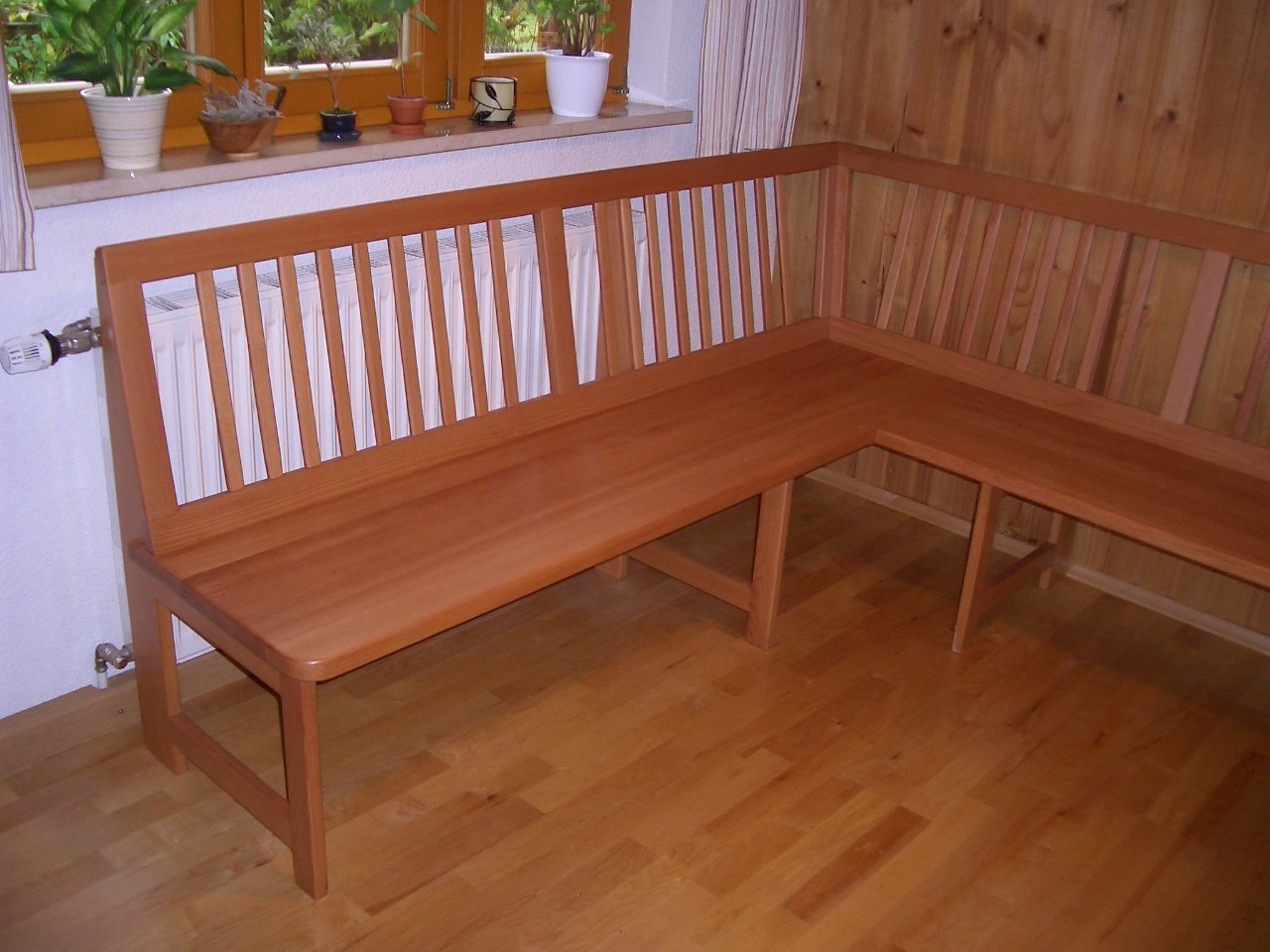 Wooden L-shaped corner bench in a room with hardwood floors, wooden paneled walls, and a windowsill with potted plants.