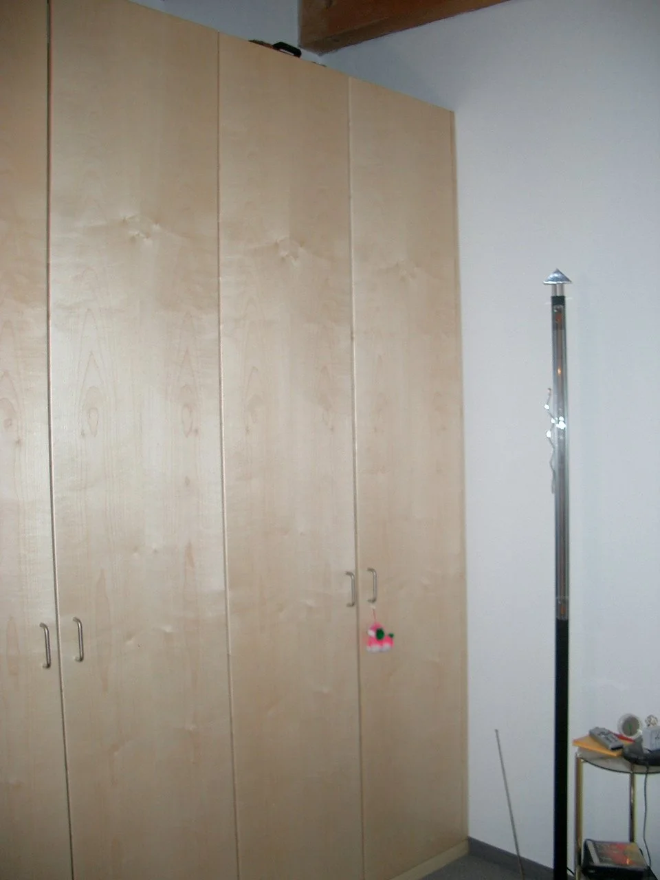 A wooden wardrobe with four doors, two of which have small handles. There is a pink and white decorative item hanging from one of the handles. To the right of the wardrobe, a tall metallic object stands beside a small table that has some items includ