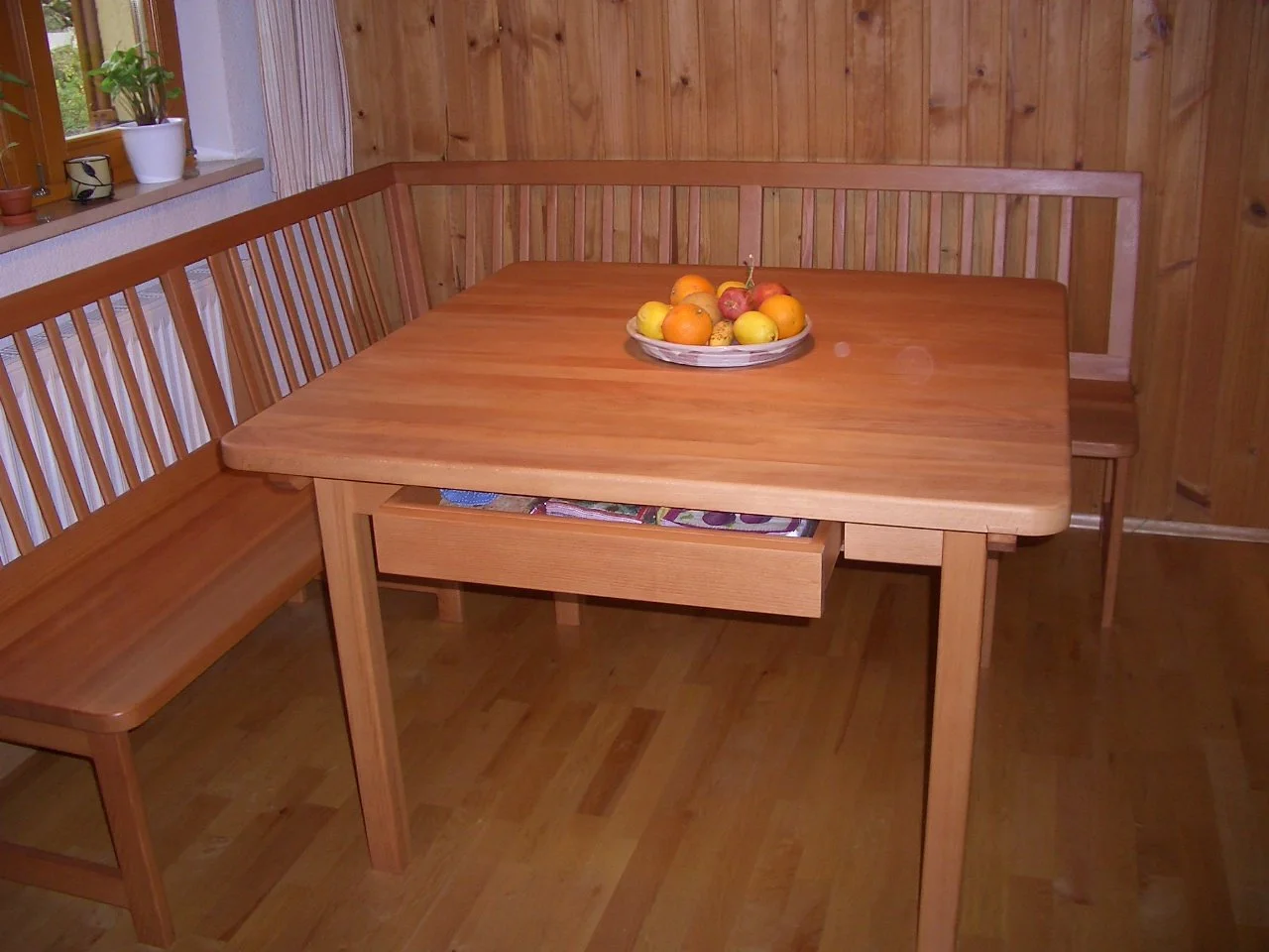 Wooden dining area with a square table, a bench along two sides, and a circular fruit bowl with apples, oranges, and lemons in the center. A window with a potted plant is visible on the left.