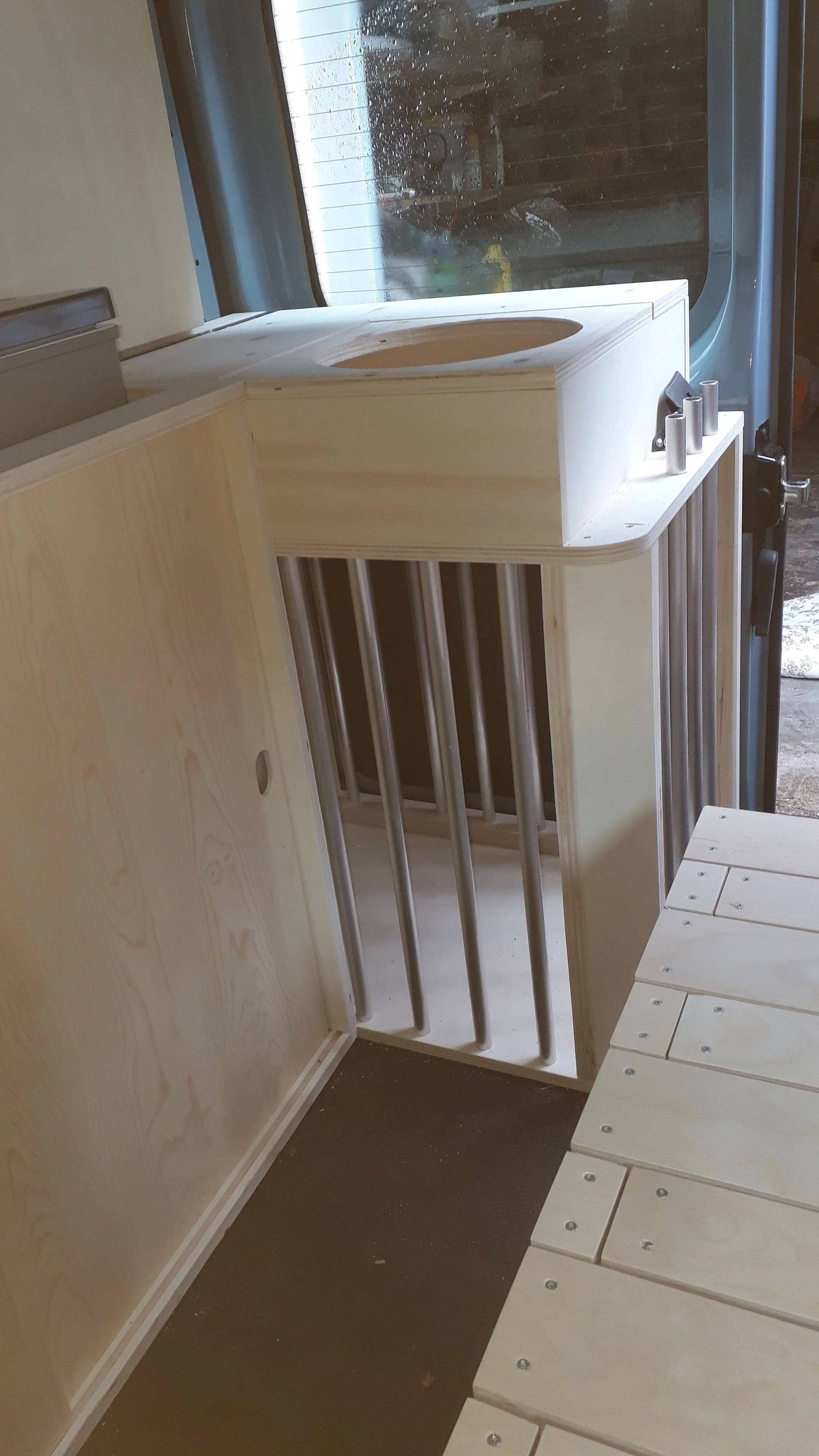 The image shows an unfinished wooden structure with vertical metal bars on the sides, likely a piece of furniture or construction. There is a window in the background with rain droplets on it, and the scene appears to be indoors near a door.