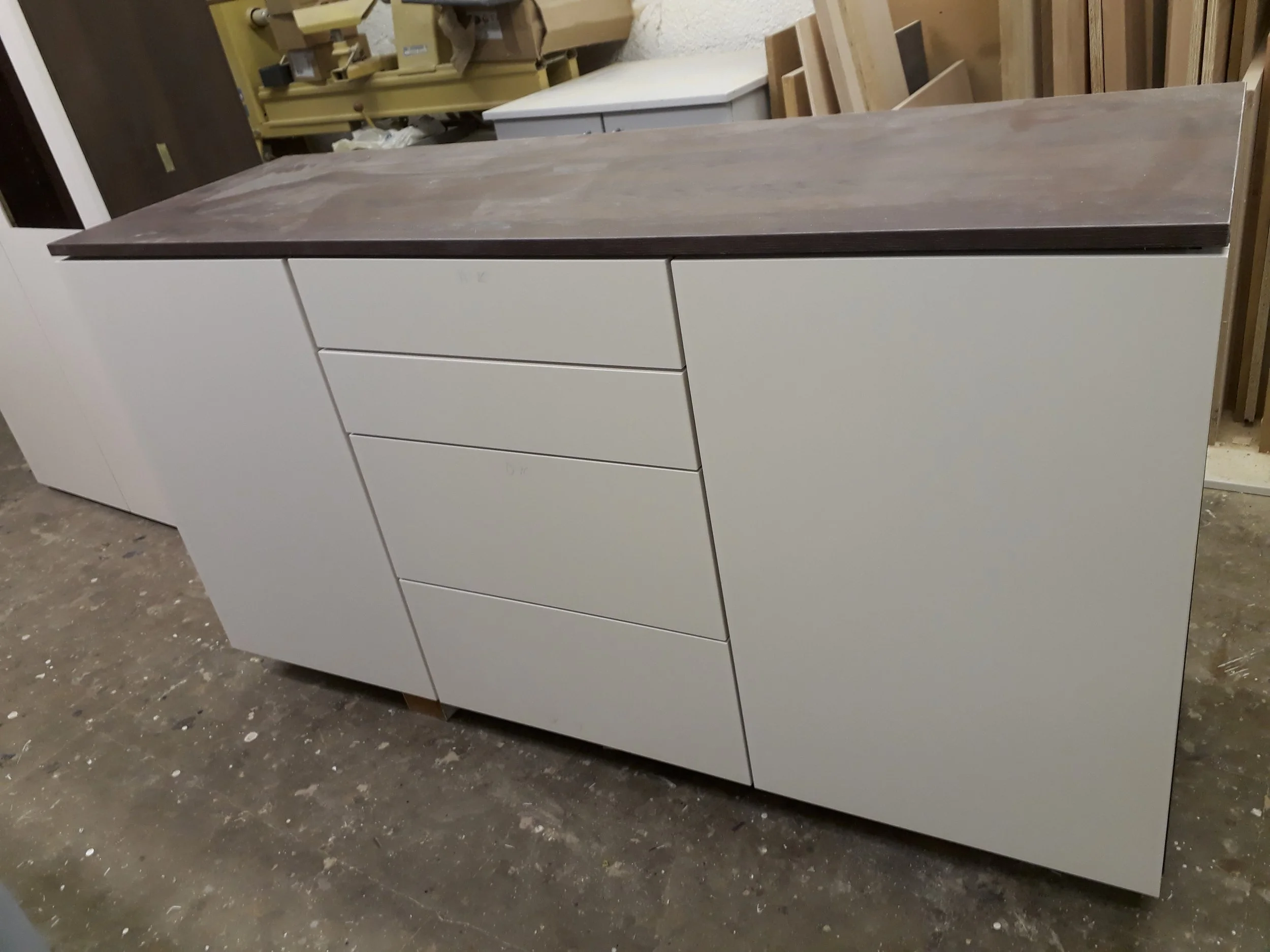 A white cabinet with multiple drawers and a dark wood top is in a workshop or warehouse setting, surrounded by wood pieces and tools.