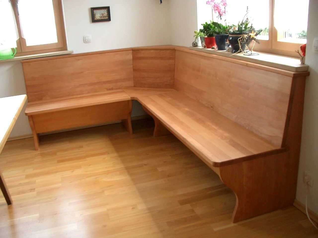 A corner wooden bench with a backrest in a room near a window, decorated with potted plants on the windowsill and a small picture on the wall.