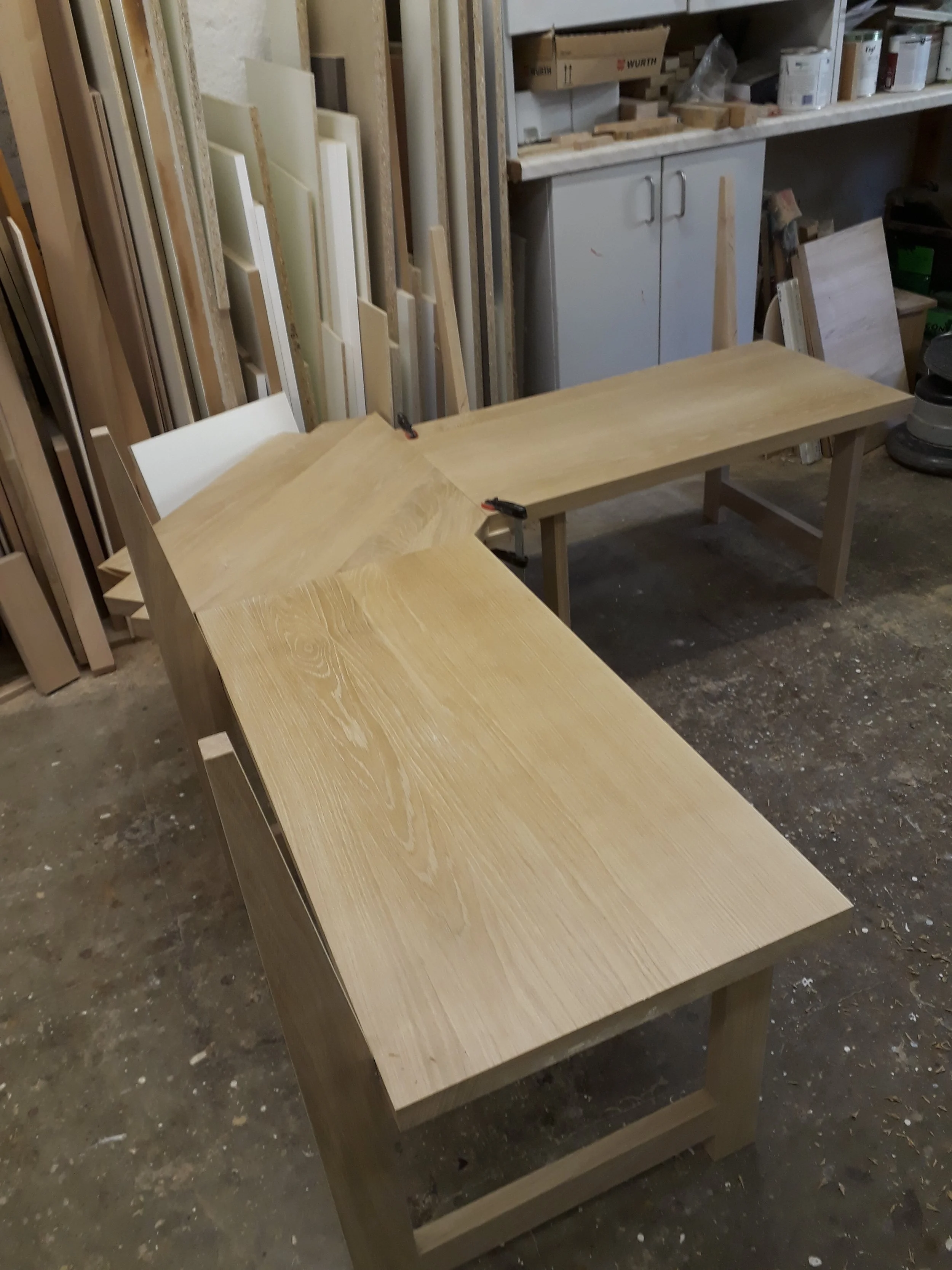 A woodworking workshop with a large wooden table in the center, clamps attached to the edge, and storage shelves filled with wood pieces, paint cans, and tools in the background.