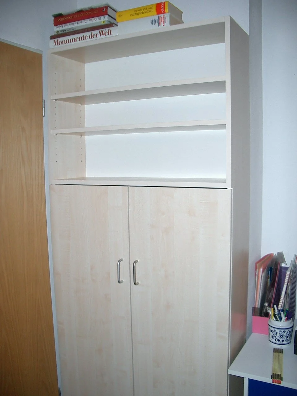 A tall white bookshelf with four empty shelves and a closed cabinet at the bottom, located next to a wooden door. Books and supplies are on top of the bookshelf and on a nearby white table.