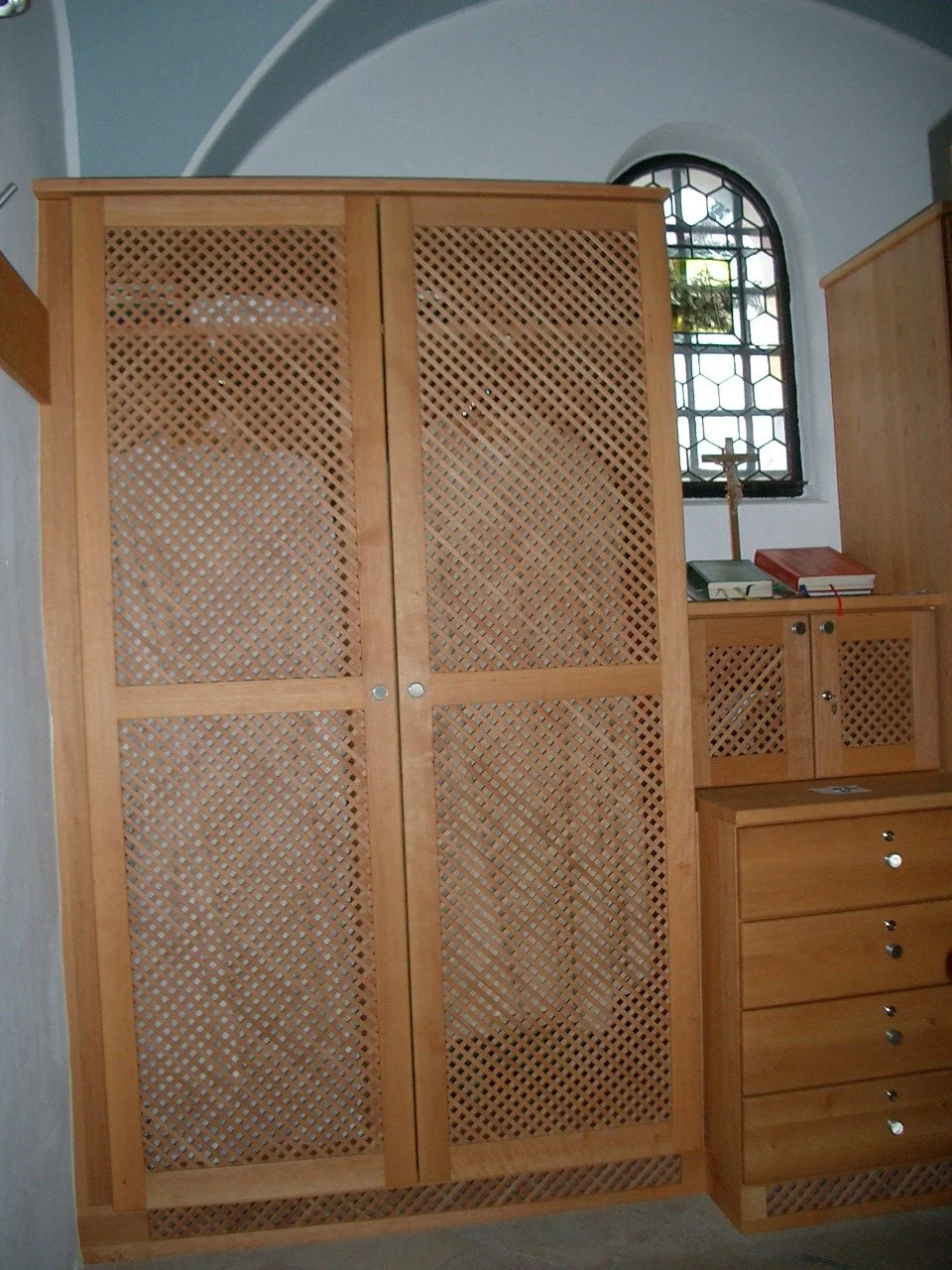 A wooden wardrobe with a lattice design, situated in a room with a window and a wooden dresser with drawers and small cabinets.