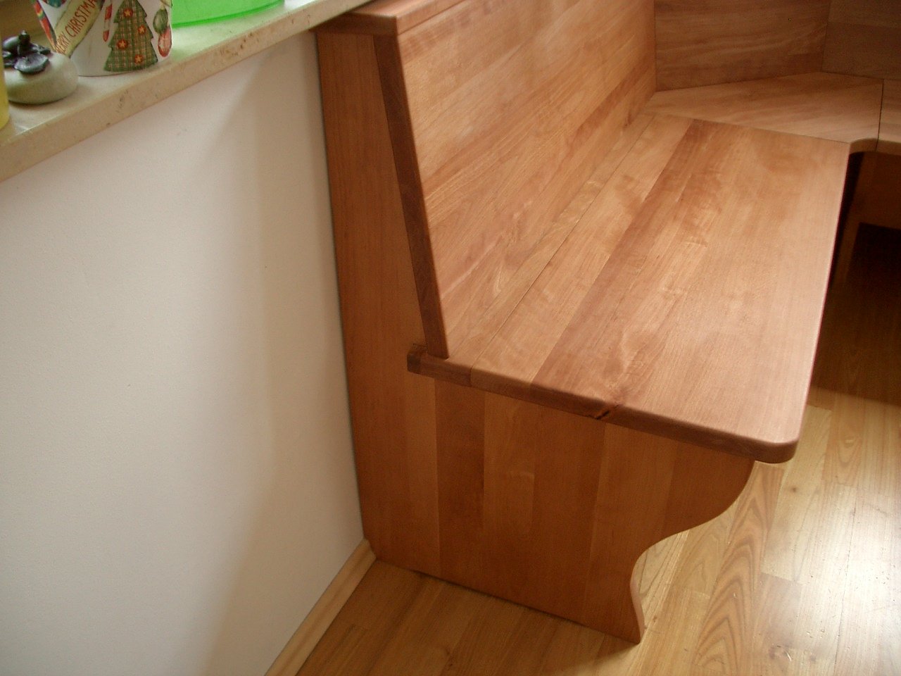 A wooden bench with a rounded edge in a room with hardwood flooring and a white wall. The bench is positioned next to a kitchen counter decorated with holiday-themed items.