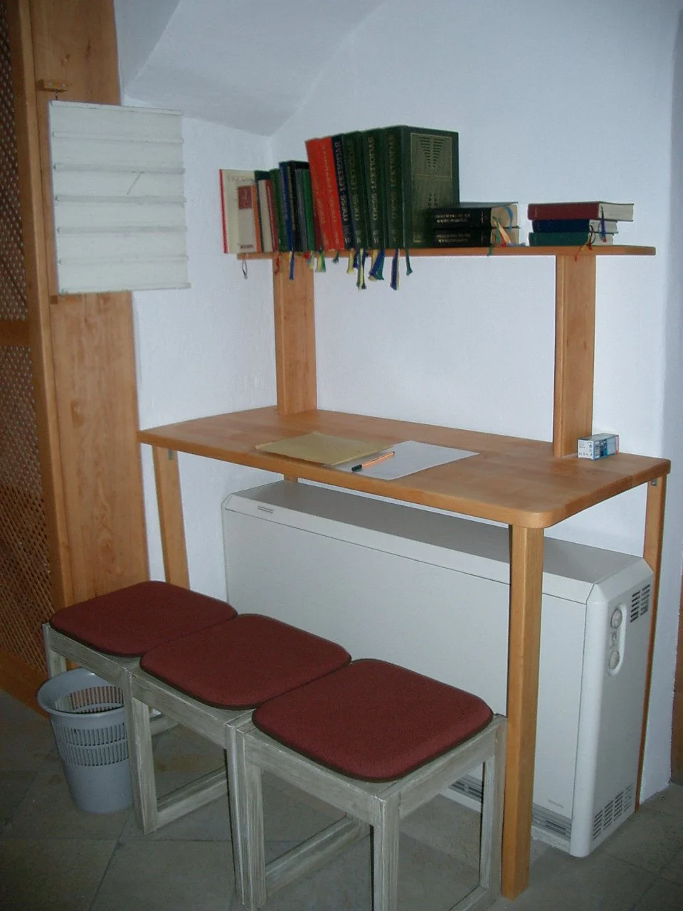 A small wooden desk with a shelf above, holding books and a notebook. Underneath the desk are three wooden stools with red cushions. To the right of the desk is a laundry dryer, and to the left is a wastebasket. The room has white walls and a tiled f