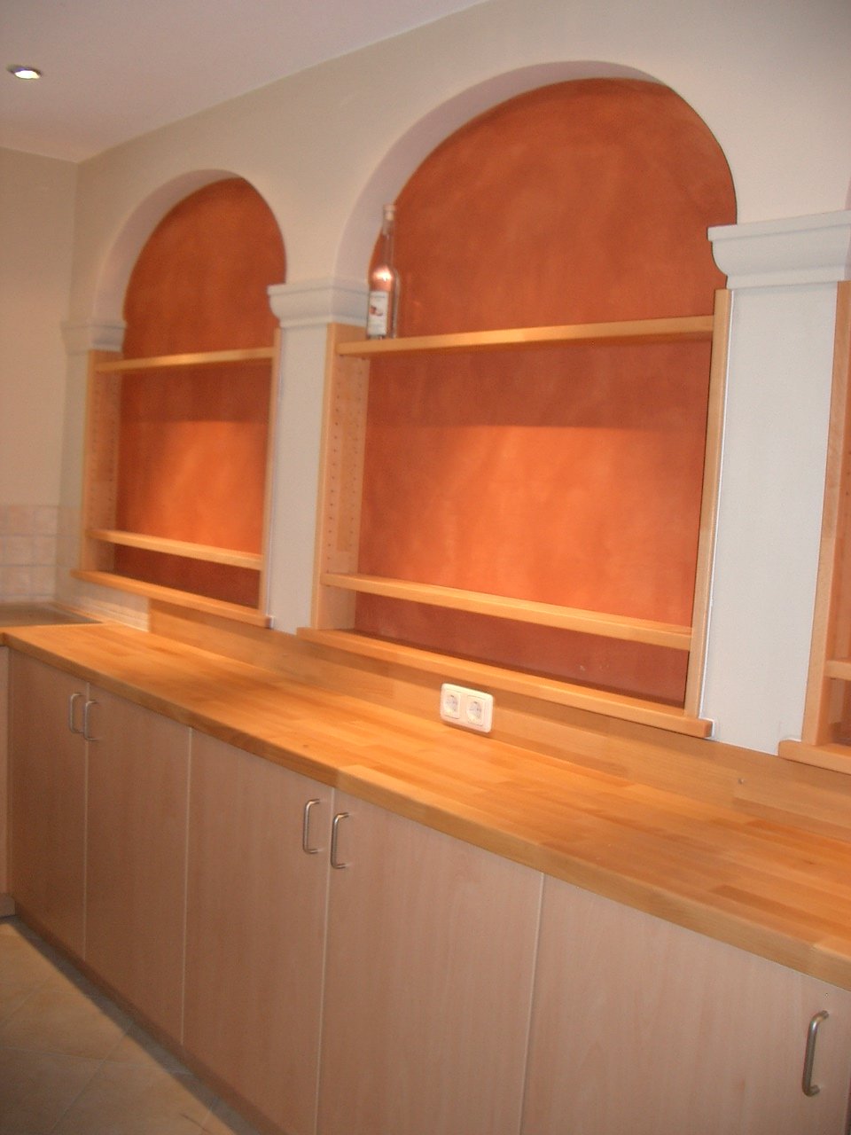 Empty kitchen with wooden cabinets, orange textured backsplash, and open shelving above a wooden countertop, with a bottle on the top shelf.
