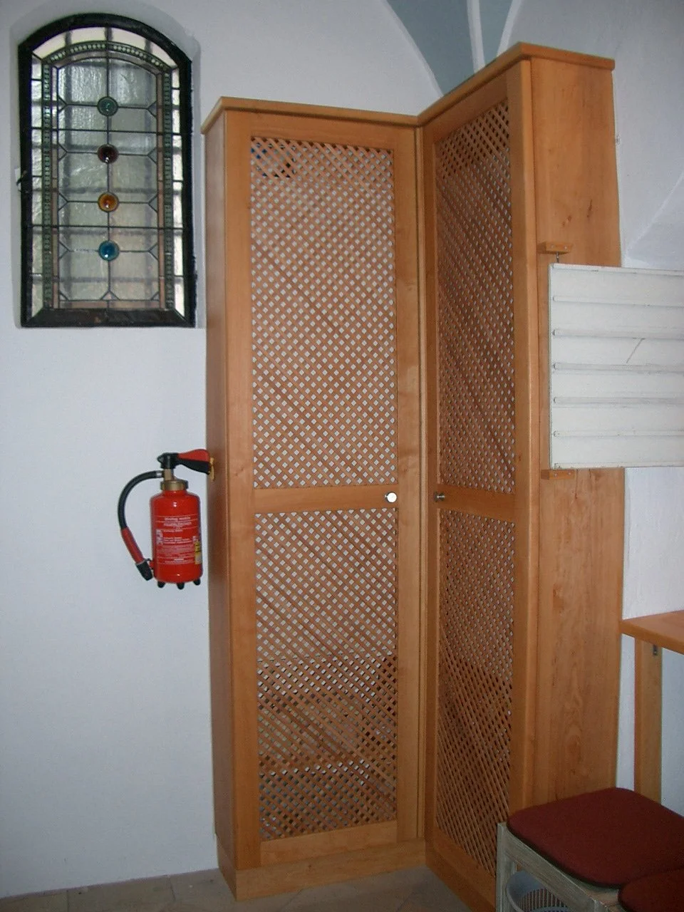 Wooden lattice room divider next to a wall with stained glass window, red fire extinguisher on the wall, and part of a wooden chair with red cushion visible.