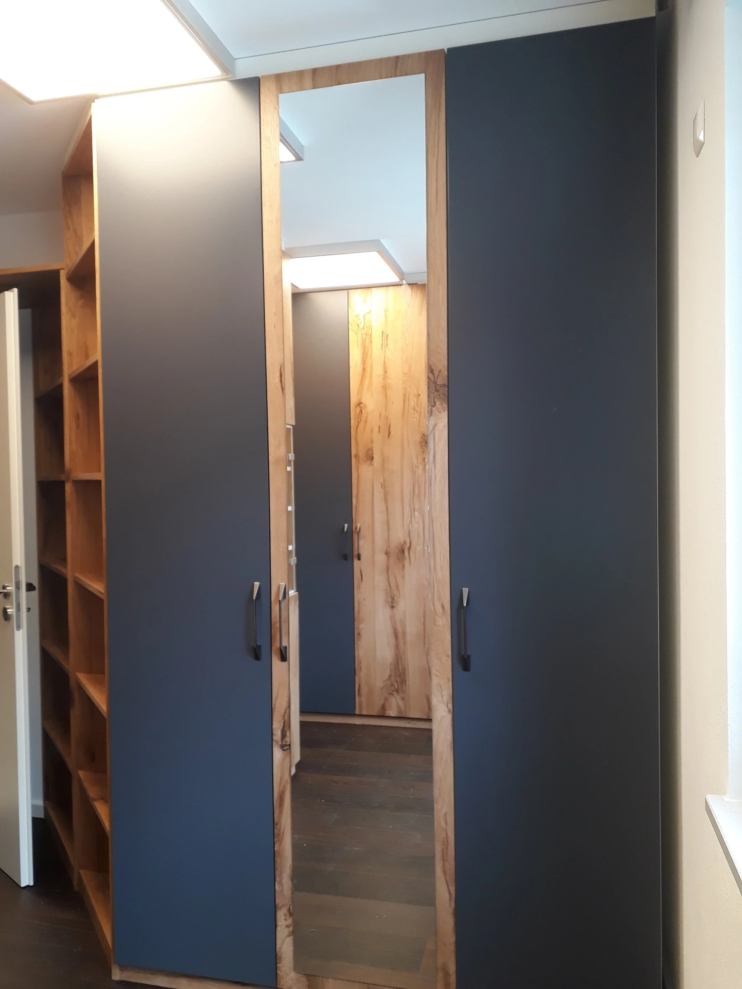 A modern room with dark blue and wooden cabinets, including a mirror, with built-in shelves and handles, and a partial view of a door with a handle.