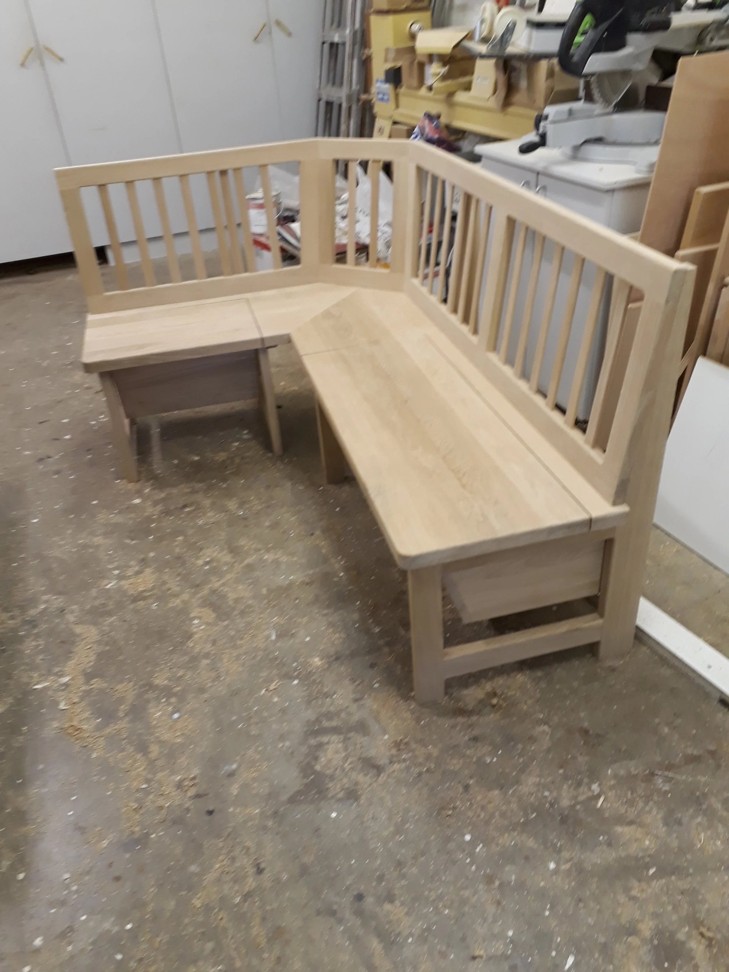 Unfinished wooden corner bench with a backrest, in a workshop or storage area with tools and supplies in the background.