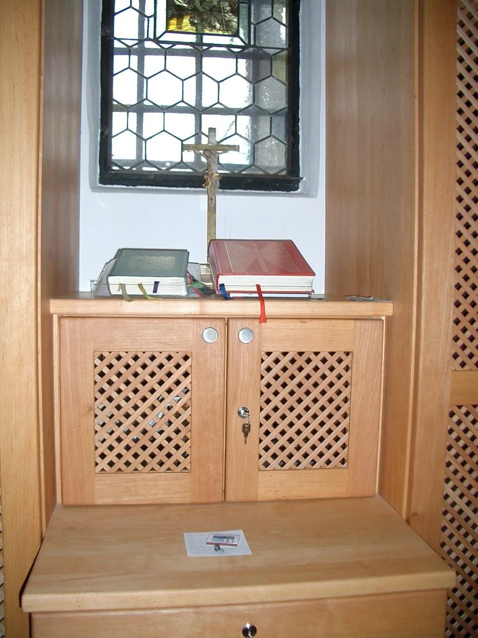 Wooden church altar with a wooden cross, Bible, candles, and a latch. Behind the altar is a window with decorative black wrought iron bars.
