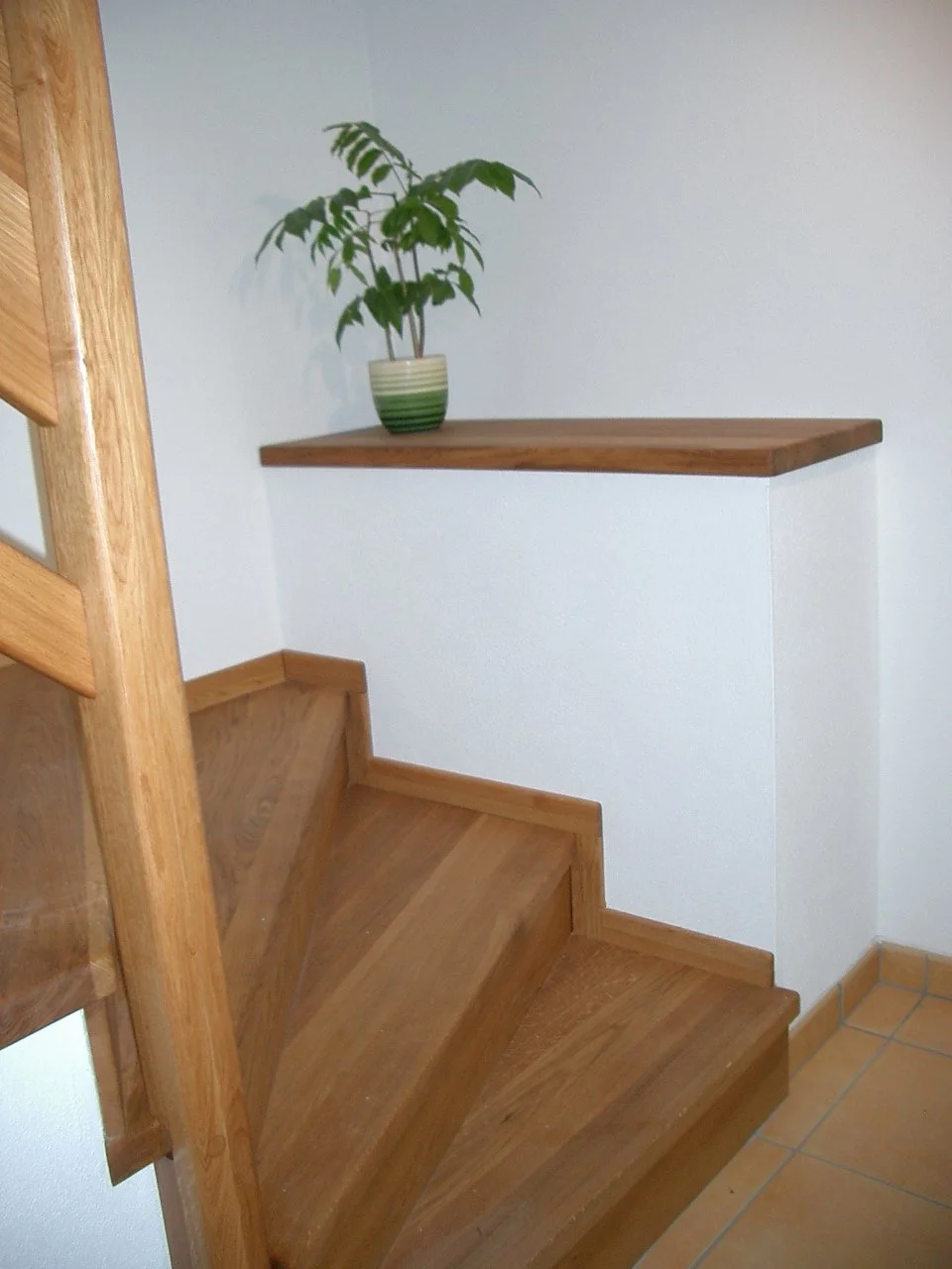 Wooden staircase with a small ledge holding a potted plant in a green and cream striped pot, against white walls.