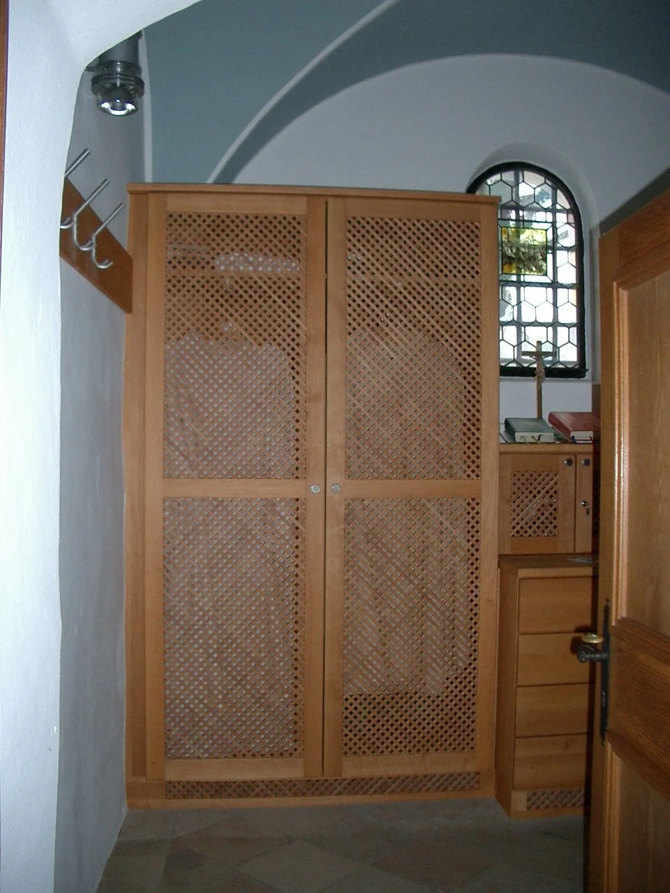 Wooden lattice cabinet with closed doors located in a room with arched ceiling, small decorative window, and wooden furnishings.