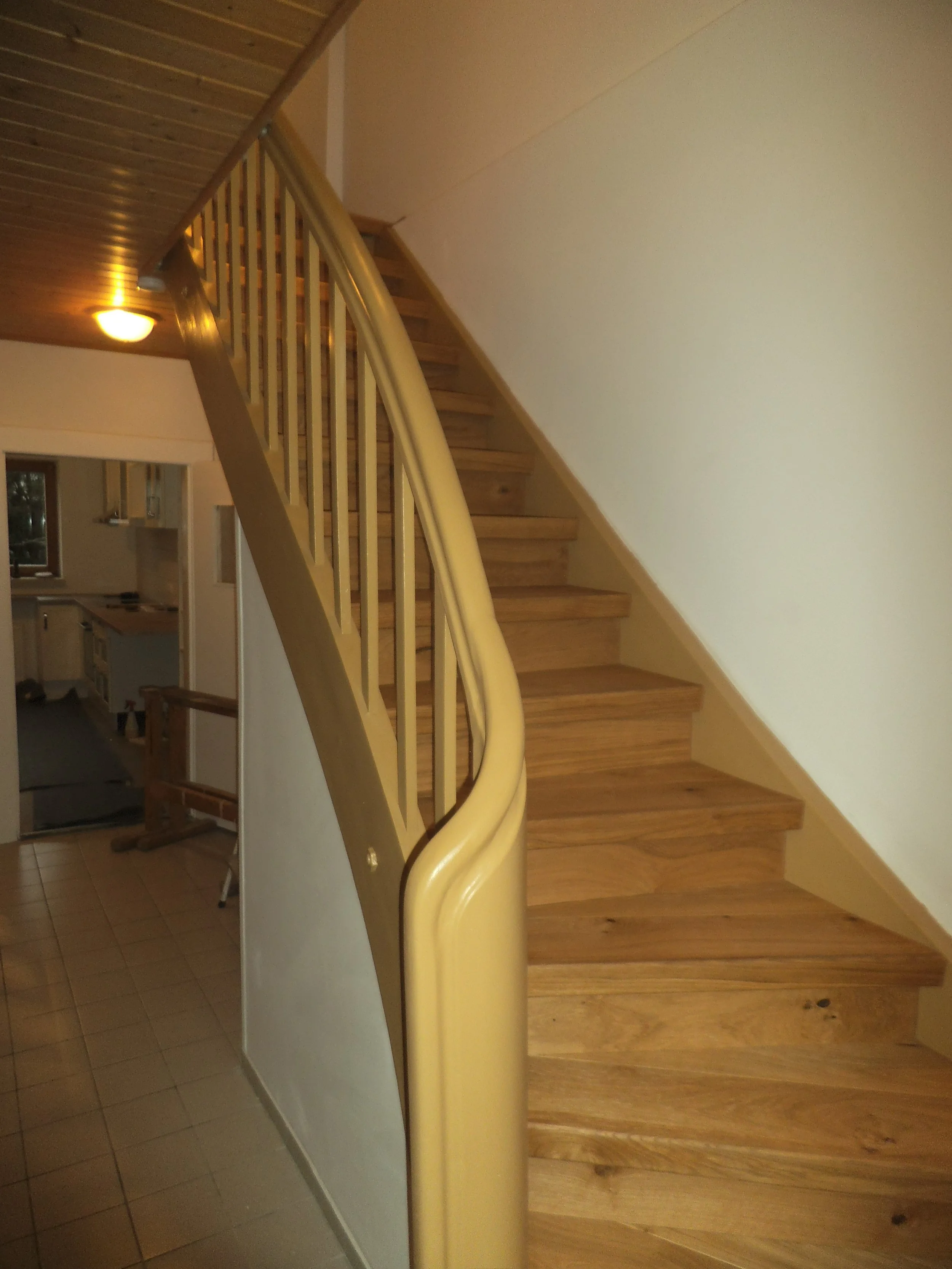 Interior view of a staircase with wooden steps and a curved yellow handrail, next to a large mirror reflecting part of the room, featuring wood-paneled ceiling and white walls.