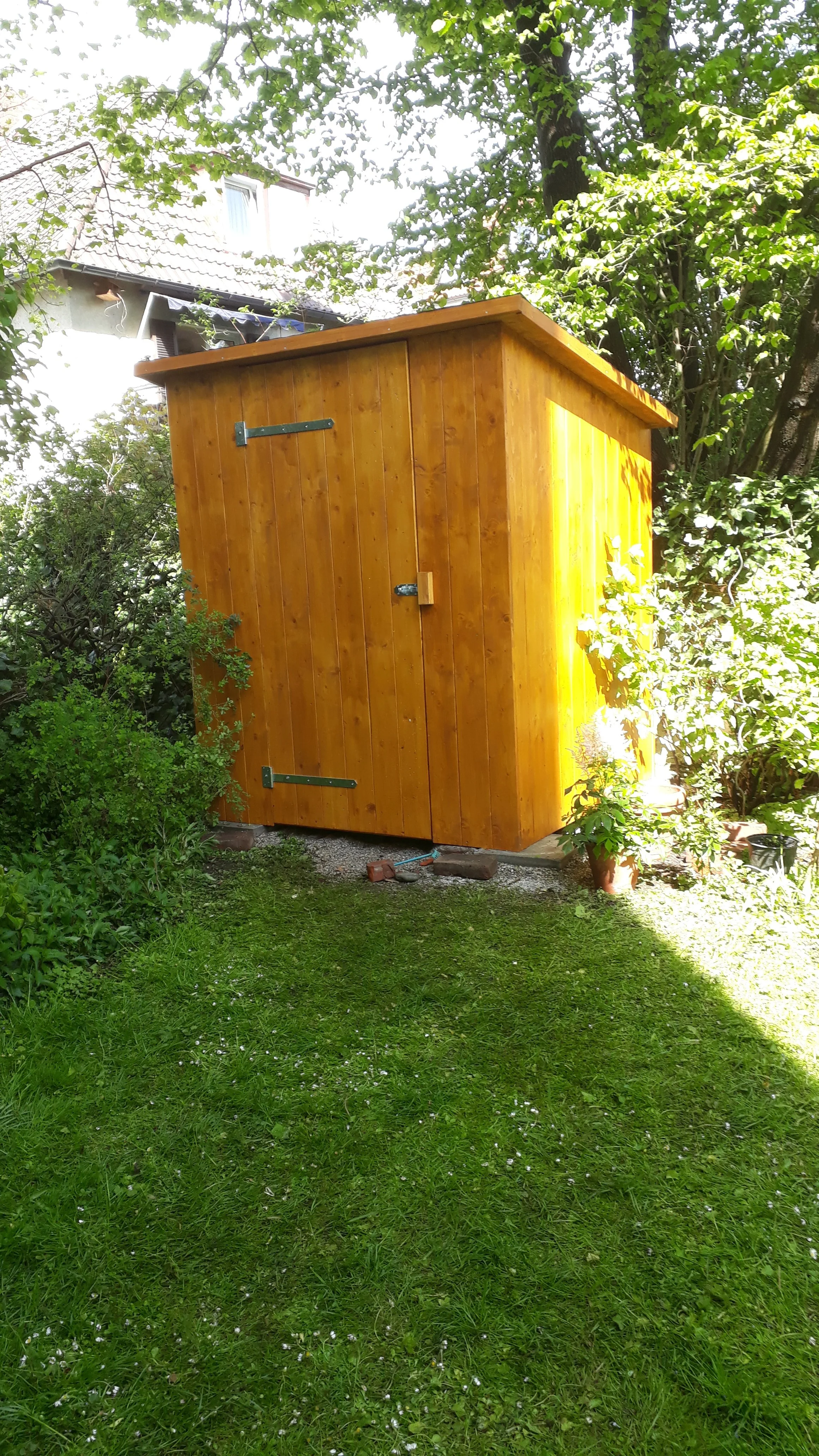 A wooden garden shed on a grassy lawn surrounded by trees and plants.