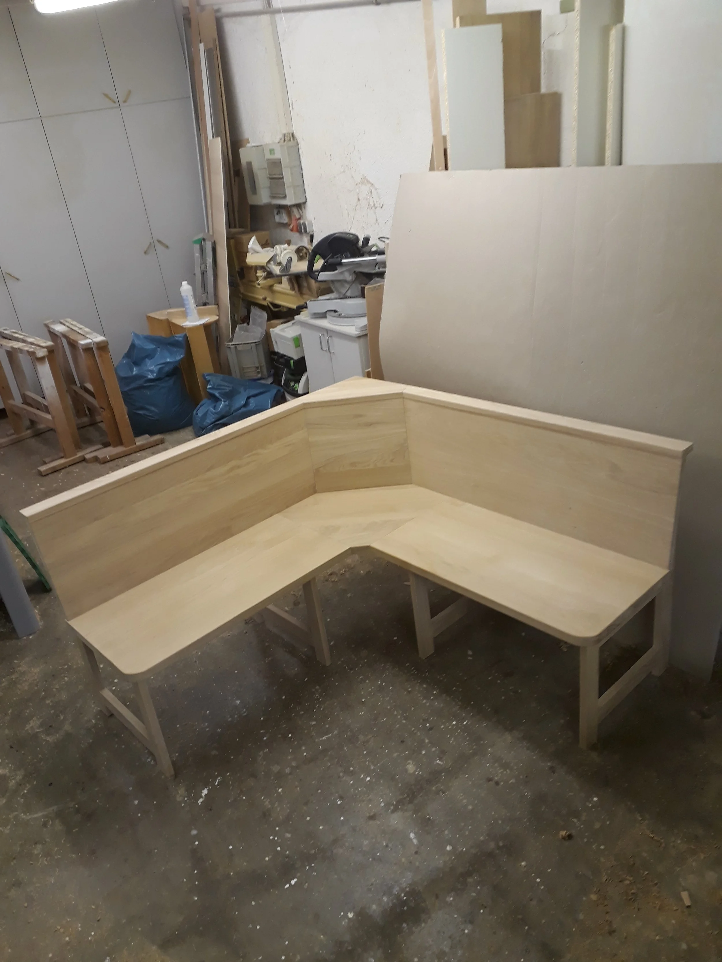 Wooden L-shaped bench in a workshop with construction tools and materials in the background.