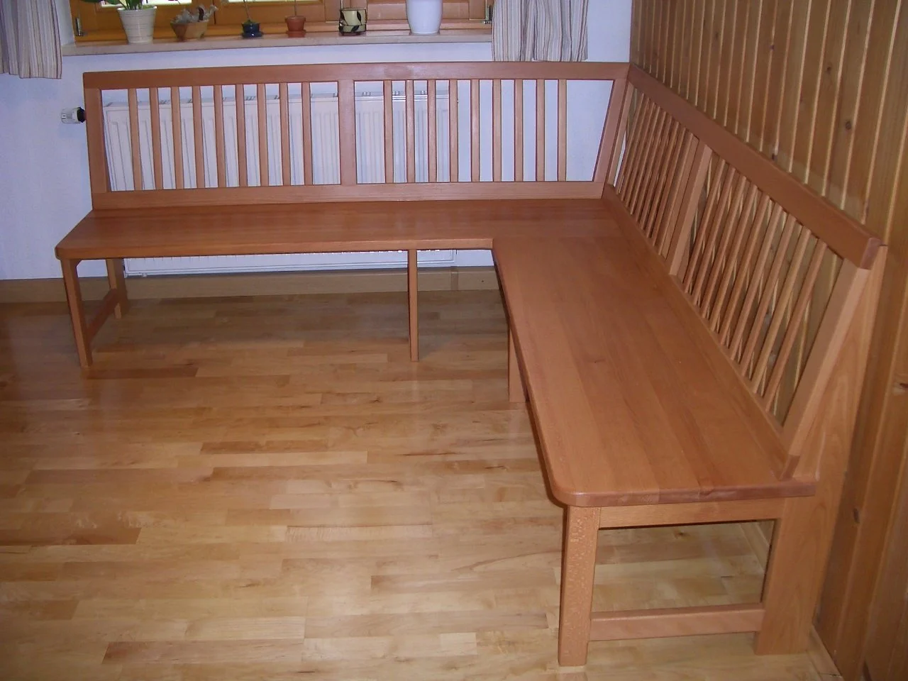 Wooden L-shaped corner bench with slatted backrest in a room with wooden floor and window with potted plants.