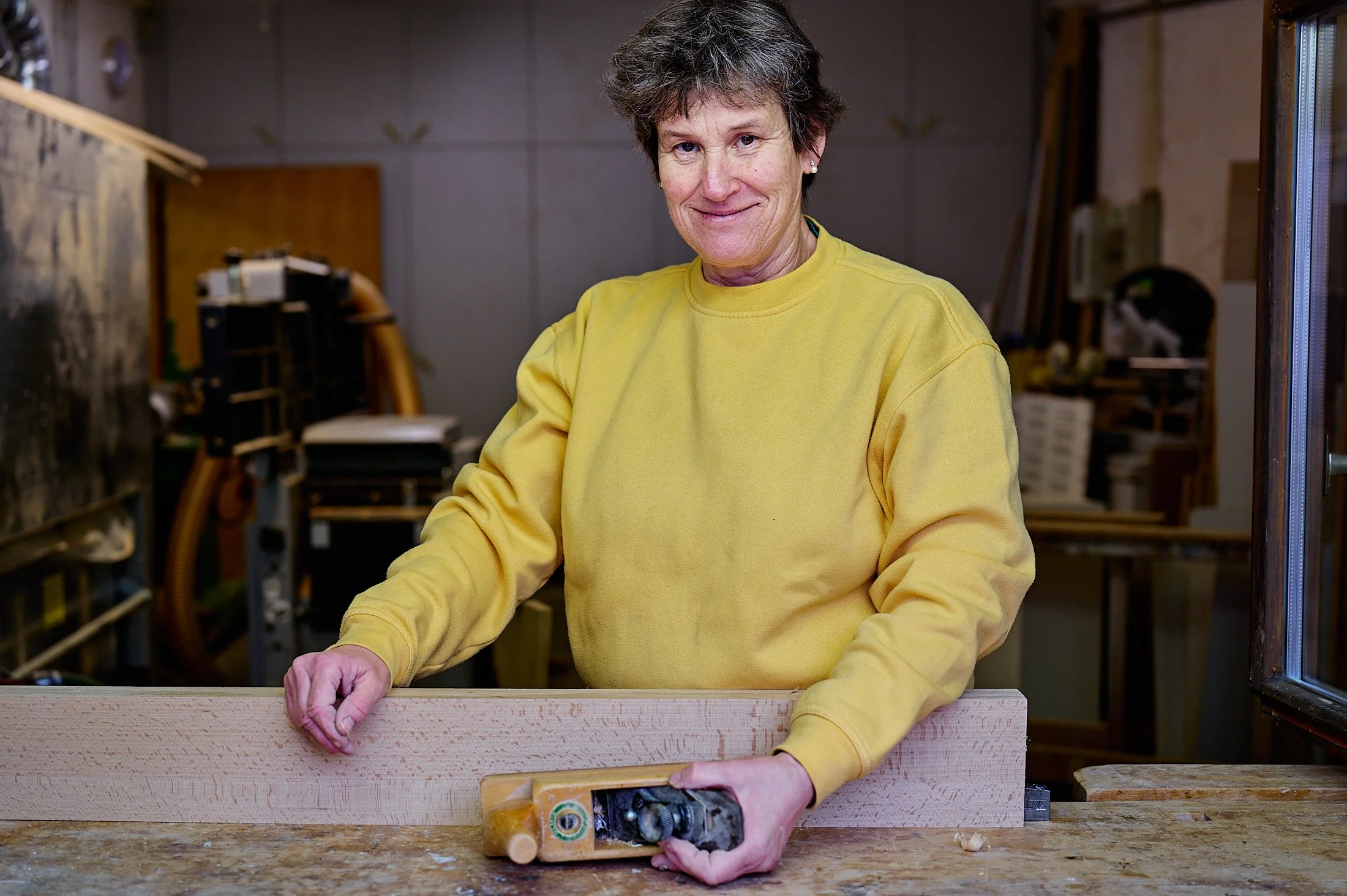 A woman with short gray hair wearing a yellow sweatshirt working in a woodworking shop, smoothing a piece of wood with a hand plane.