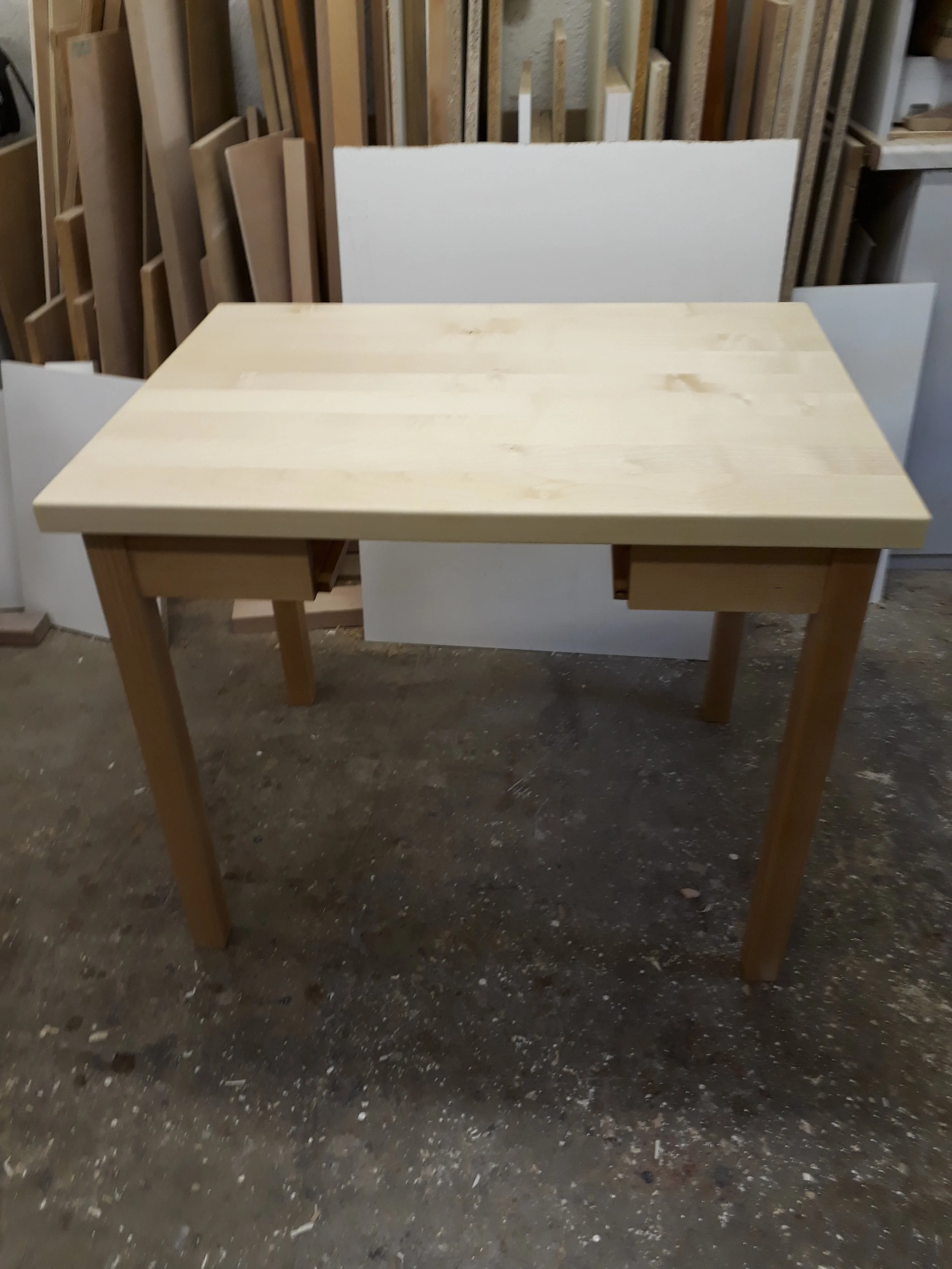 A wooden table with a light natural finish, standing on a concrete floor, with several woodworking panels and boards leaning against the wall behind it in a workshop or storage area.