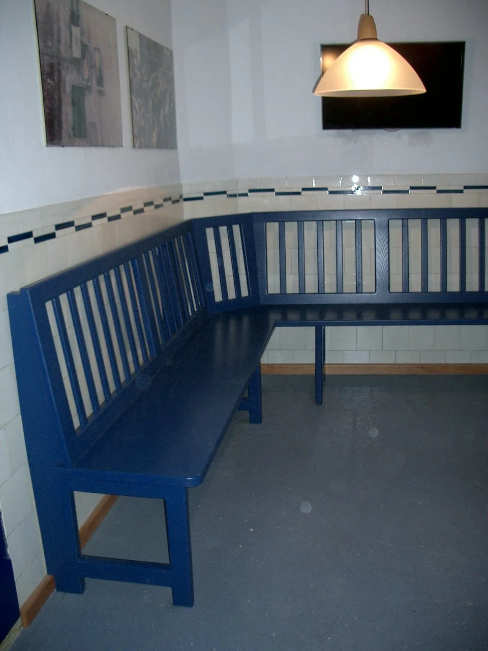 Blue metal corner bench with slatted back and seat in a room with tiled walls, a hanging lamp, and a TV mounted on the wall.