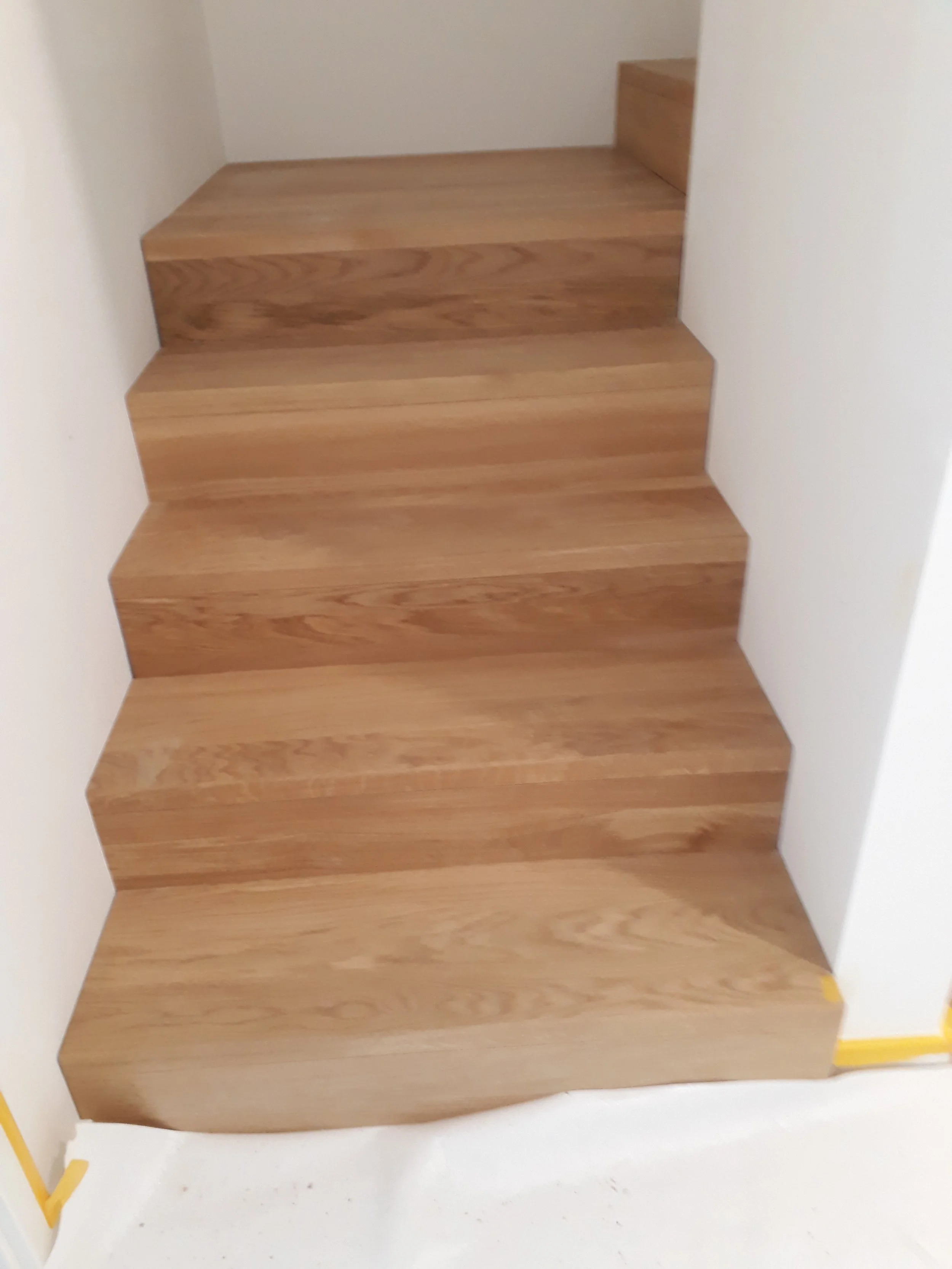 Wooden staircase in a partially finished room with white walls and protective tape along the floor edges.