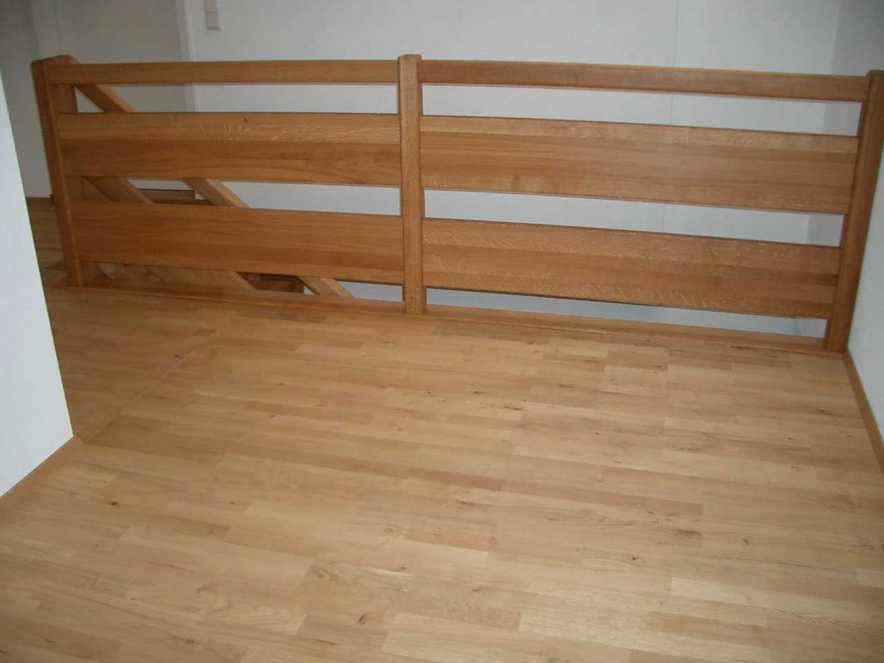 Wooden staircase railing with three horizontal slats on each side, next to a matching wooden floor and white walls.