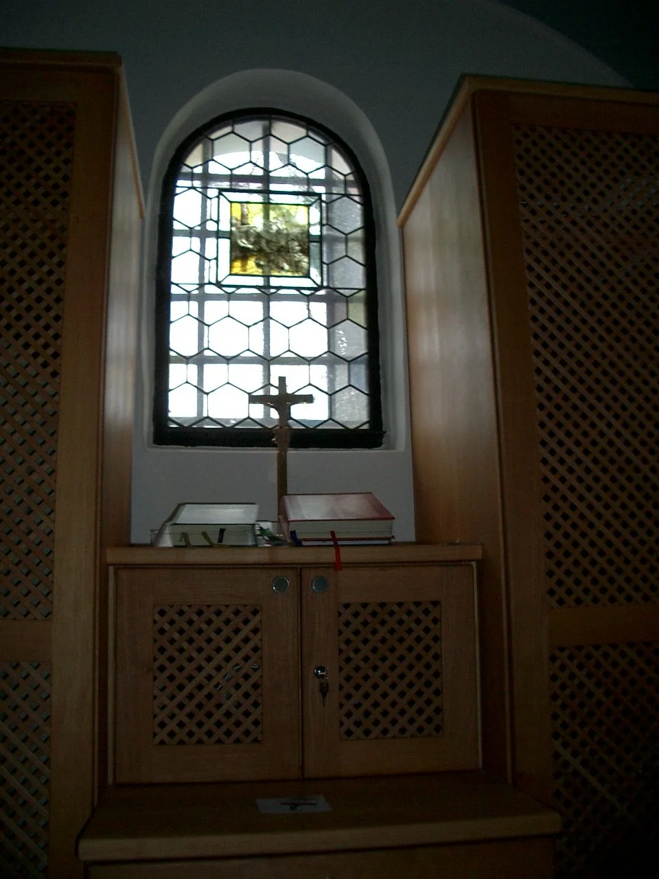 Interior view of a small prayer area with a wooden cabinet, a crucifix, prayer books, and a stained glass window.