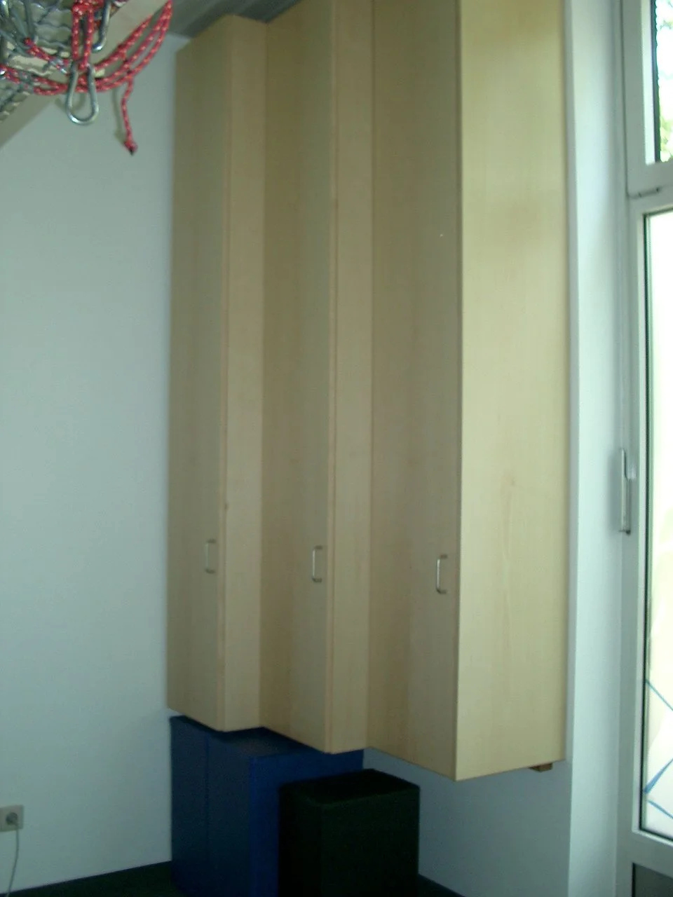 A corner of a room with wooden cabinets mounted on the wall next to a window, a small black stool beneath the cabinets, and a light-colored wall.