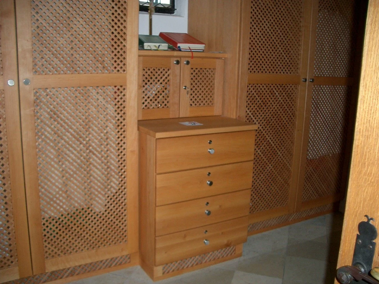 A wooden cabinet with a drawer and a small latch, set against a wall of matching wooden lattice cabinets, with a small top shelf holding books.