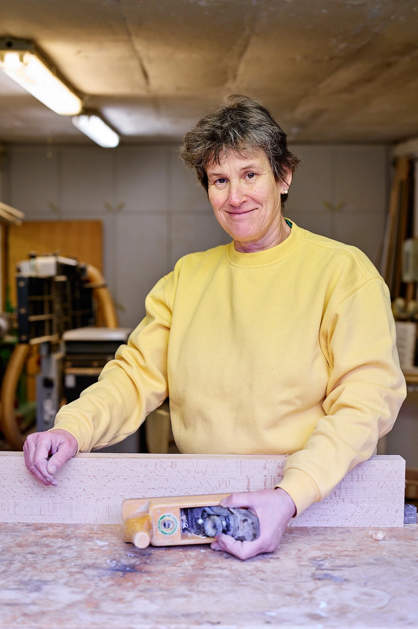 Eine Frau Sabine Zipperer Schreinermeisterin in einem gelben Sweatshirt arbeitet in einer Werkstatt an einem Holzprojekt.