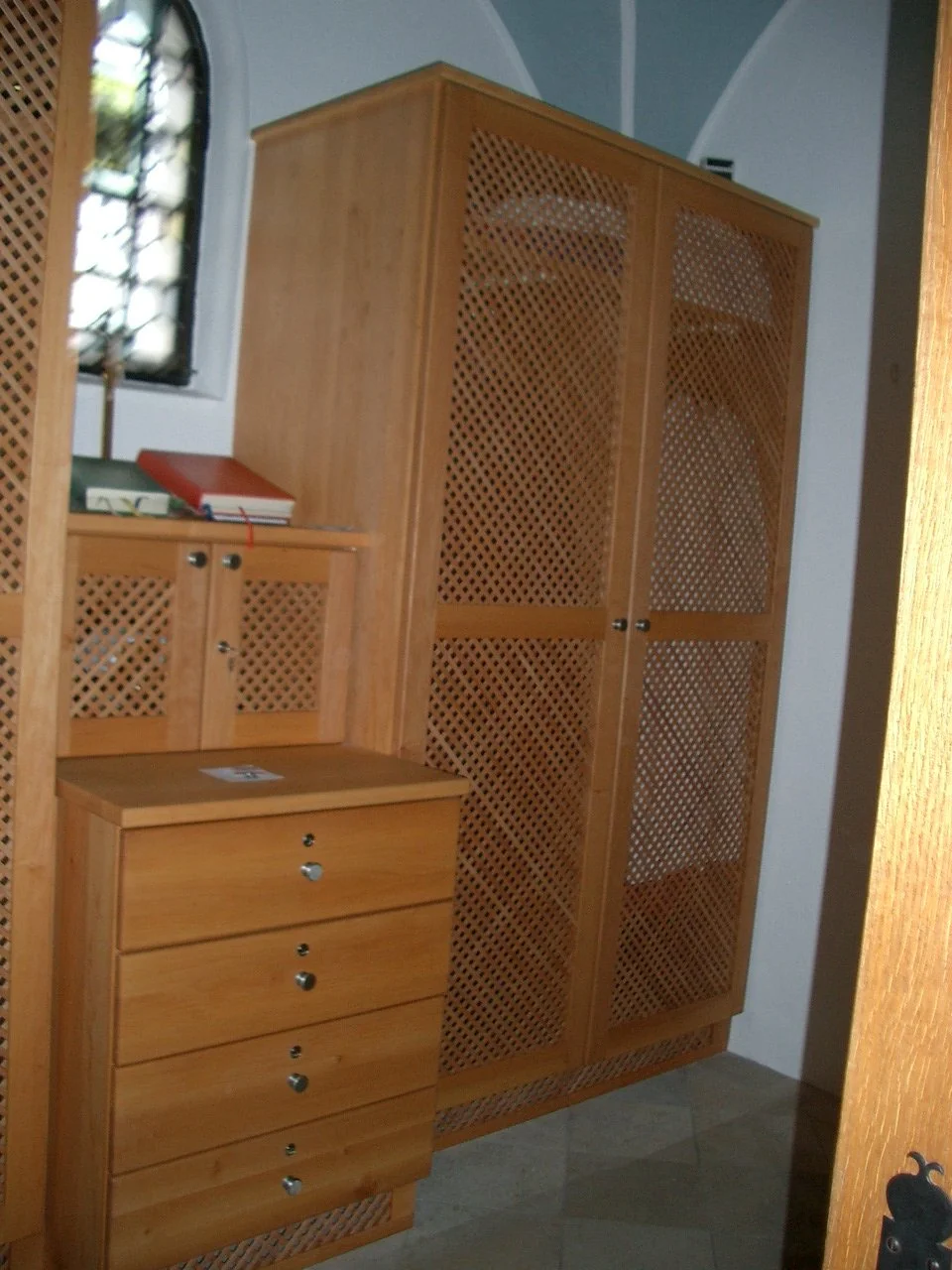 A wooden cabinet with lattice doors and a small matching chest of drawers, positioned in a corner near a window with iron bars. The cabinet contains plates visible through the lattice. The scene appears to be in a room with tiled flooring.