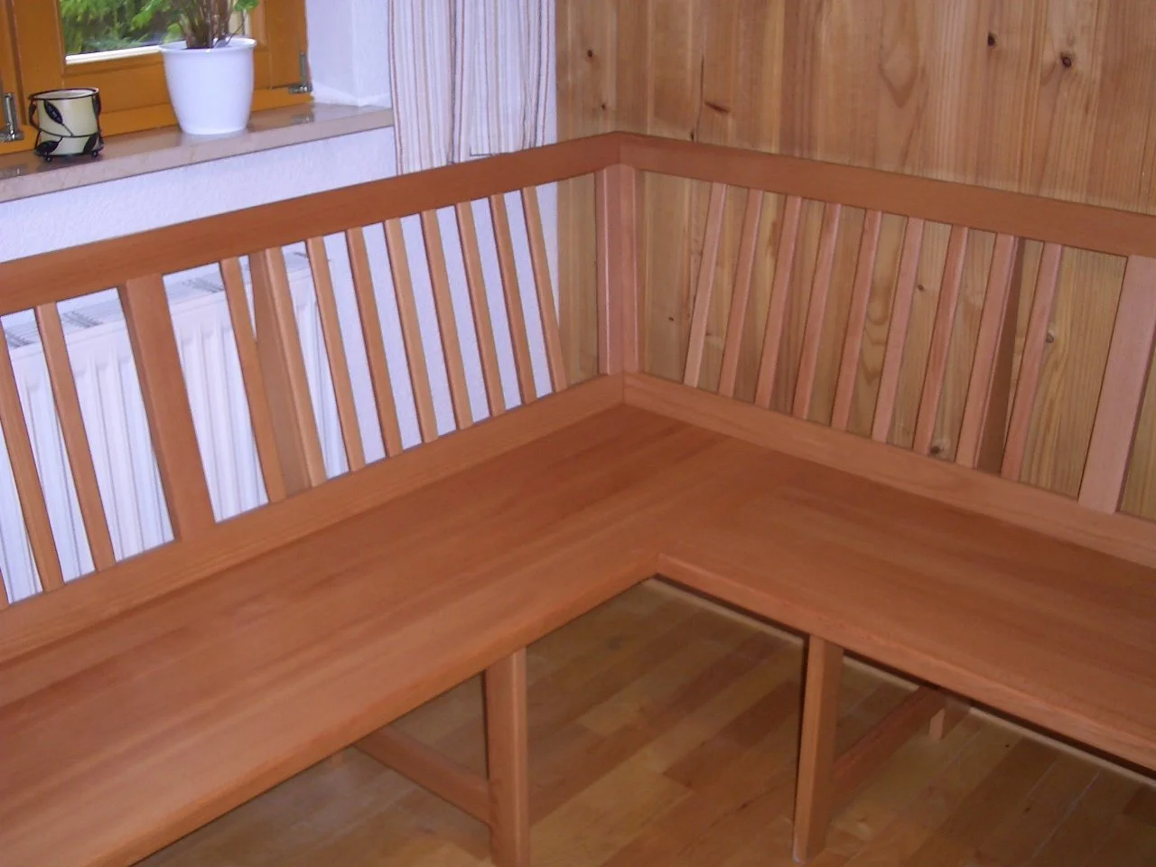 An L-shaped wooden bench with vertical slat backrest, situated in a corner near a window with a potted plant and small decorative items on the windowsill.