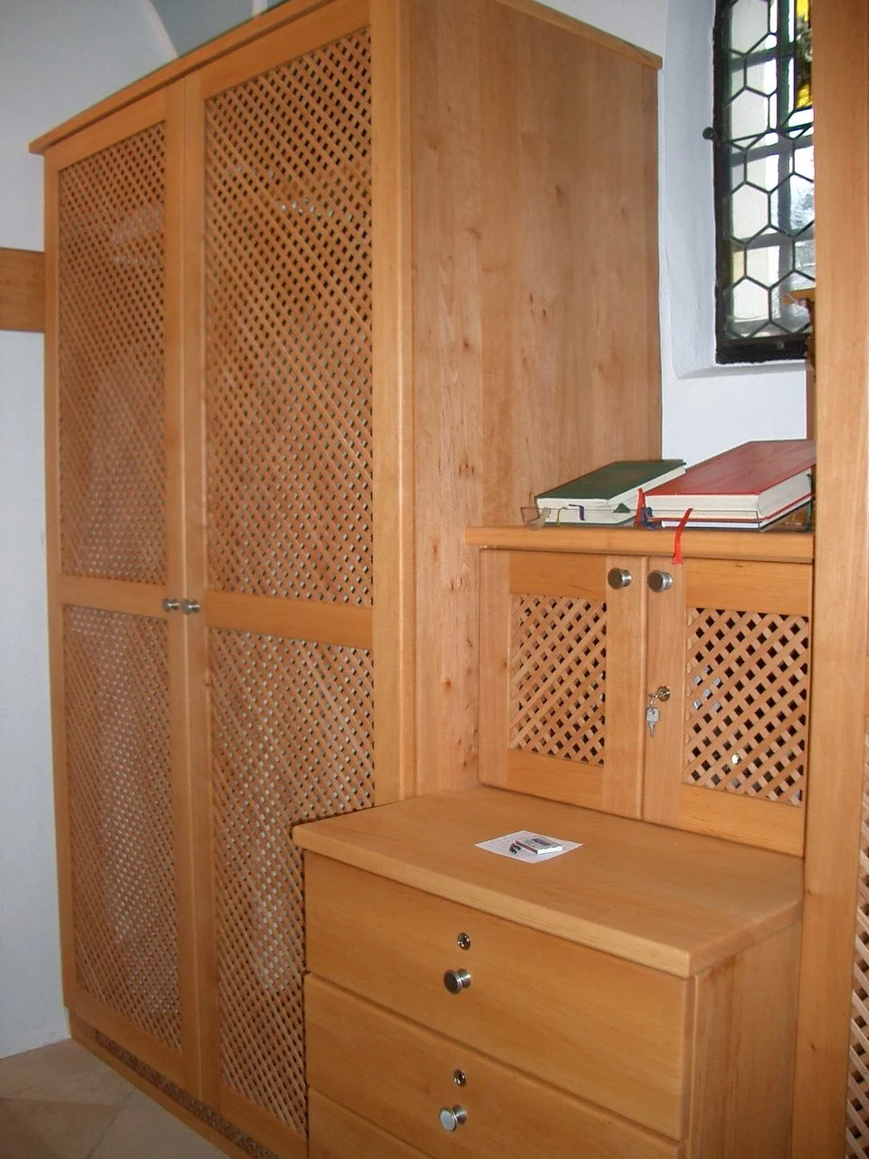 A wooden cabinet with louvered doors and drawers, placed against a wall near a window with decorative metal grille.