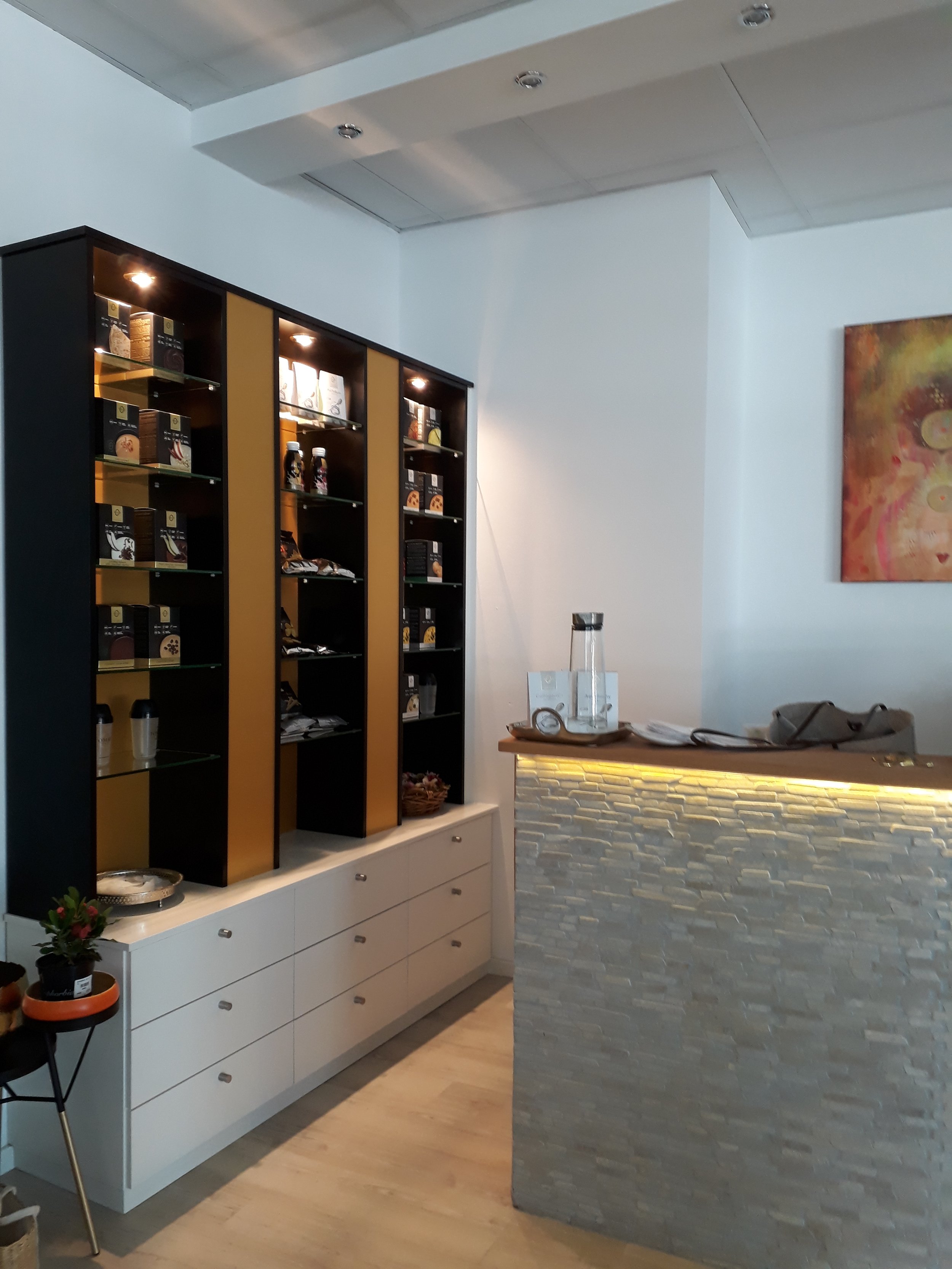 Interior of a modern coffee shop or bakery with a black and gold display shelf filled with packaged products, a white cabinet underneath, a small table with two plants, a reception counter with glassware and papers, a textured stone wall, and a color