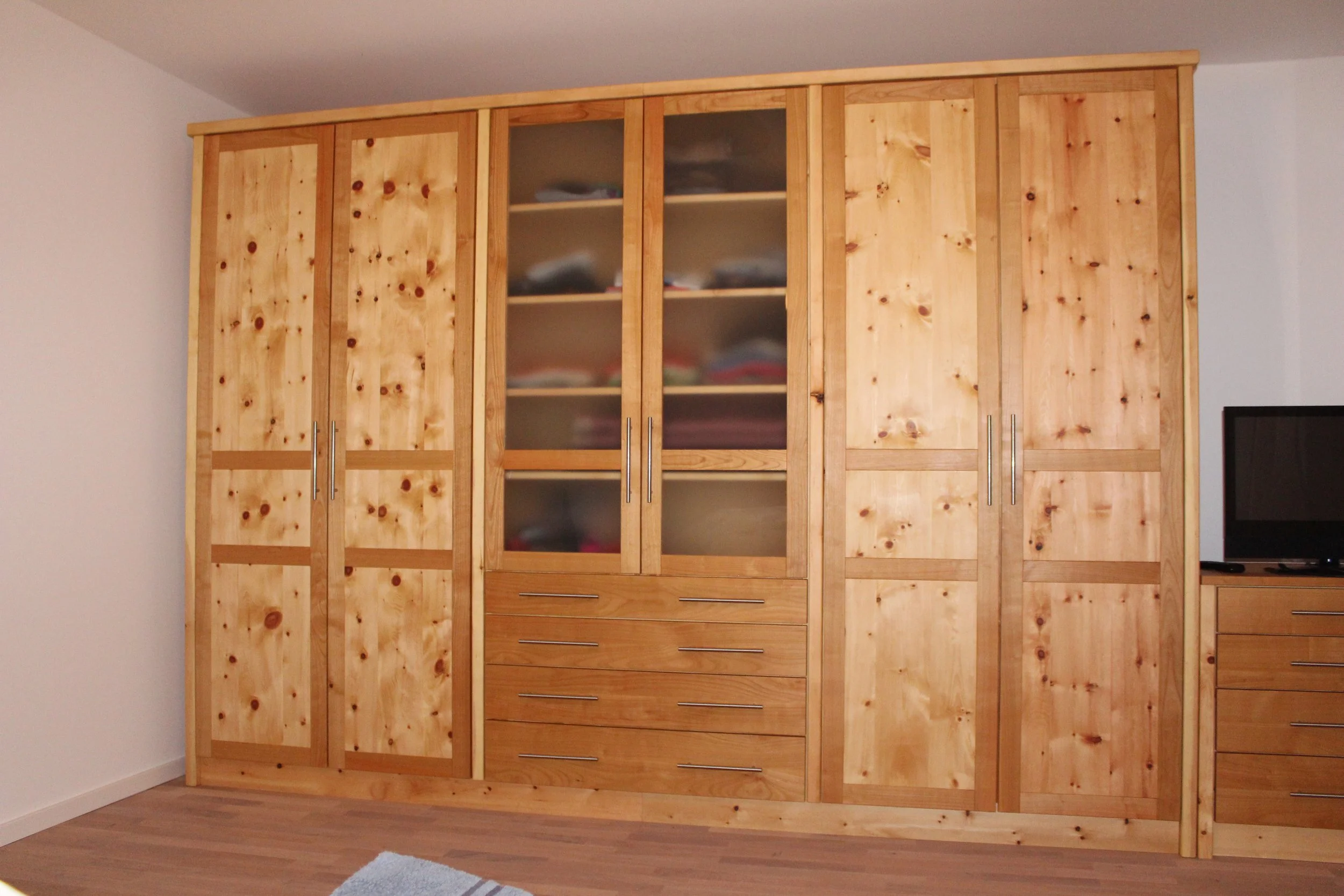 A large wooden wardrobe with glass-paneled doors and multiple drawers in a room with wooden flooring and white walls.
