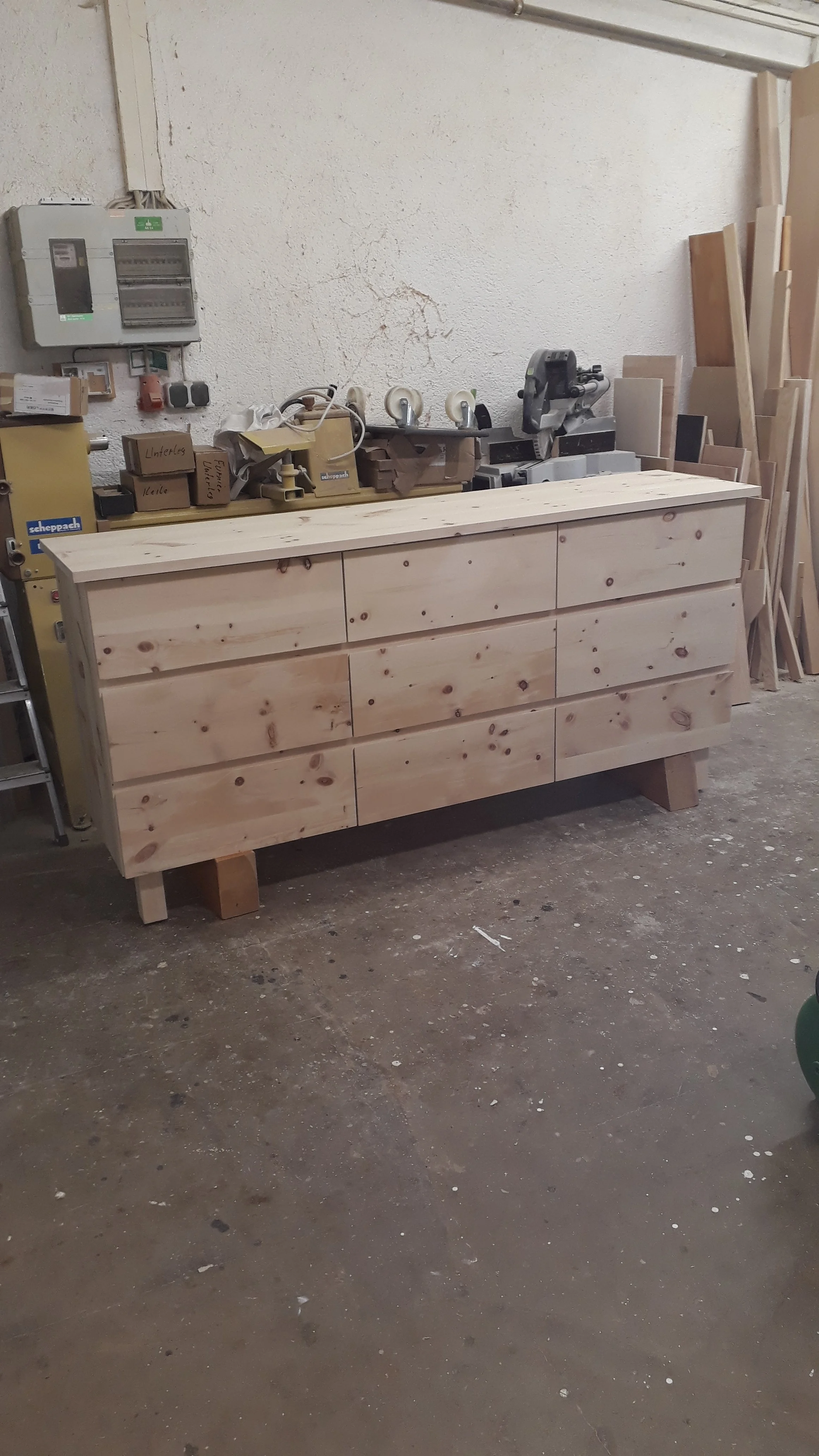 A woodworking workbench with several drawers in a workshop, surrounded by various woodworking tools and pieces of wood, against a white textured wall.