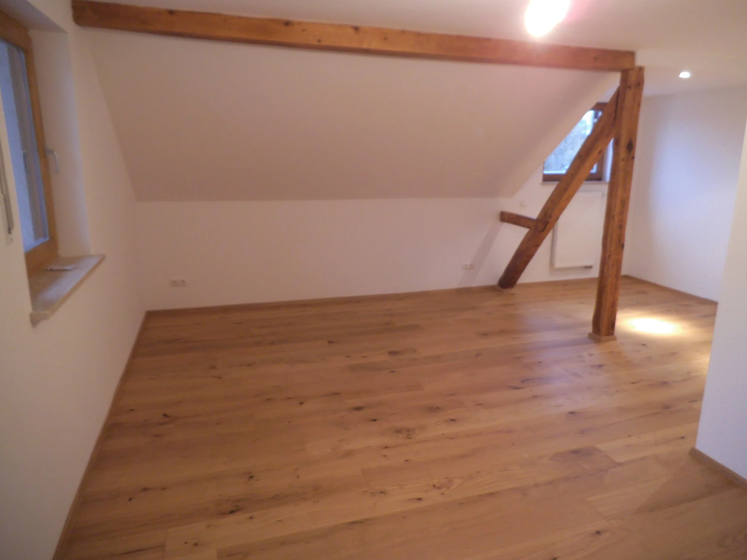 Photograph of an attic room with wooden flooring and a sloped ceiling. A small window is visible, and there is a wooden support beam across the room, with recessed lighting installed in the ceiling.