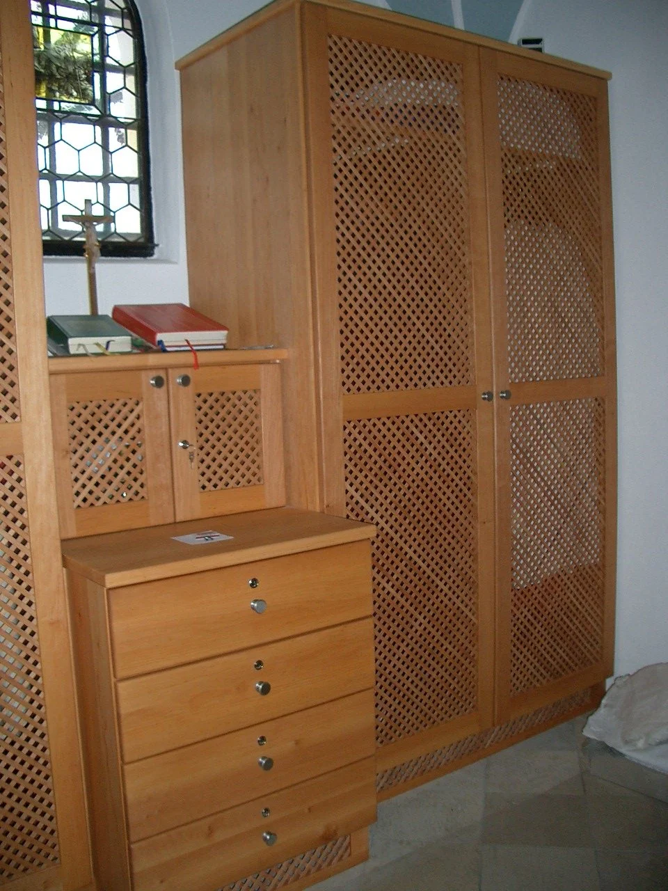 Wooden furniture with lattice doors, a small chest of drawers, a window with stained glass, and religious items including a cross and books.