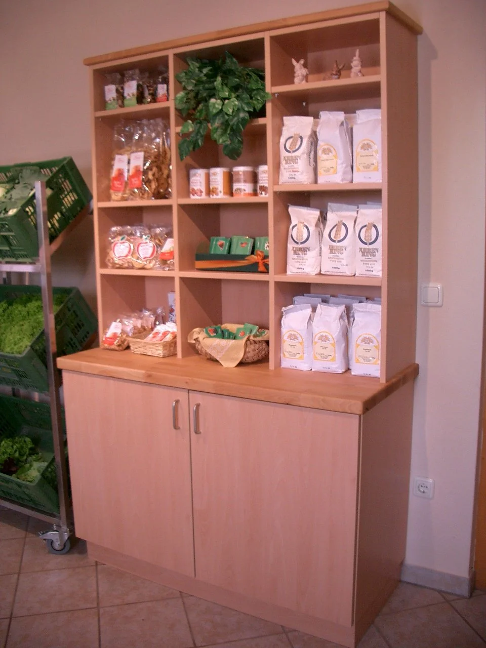 A wooden shelving unit with various packaged food products, a potted plant, and decorative items, positioned next to a metal shelf with green produce in a grocery store.