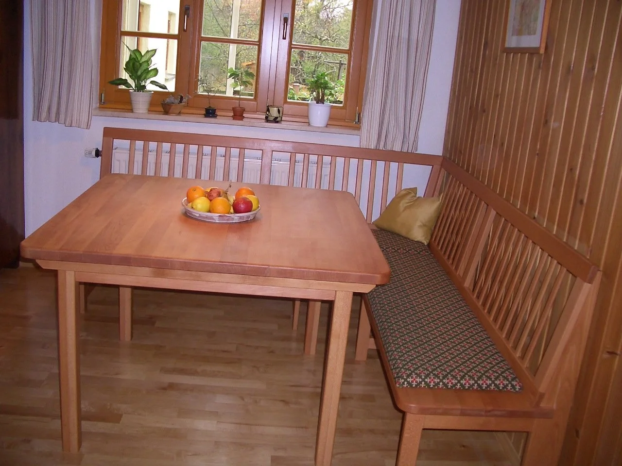 Wooden dining area with a rectangular table, a built-in bench with patterned cushion, and a window with potted plants on the sill.