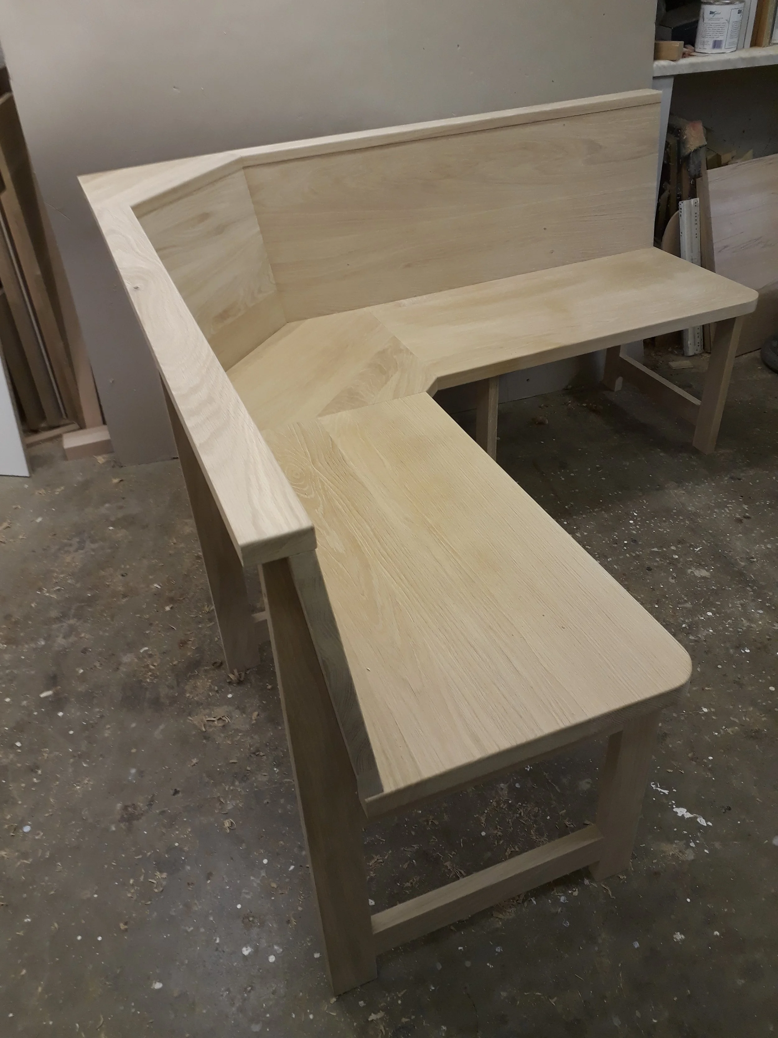 A light-colored wooden L-shaped bench in a workshop setting with shelves and woodworking supplies in the background.