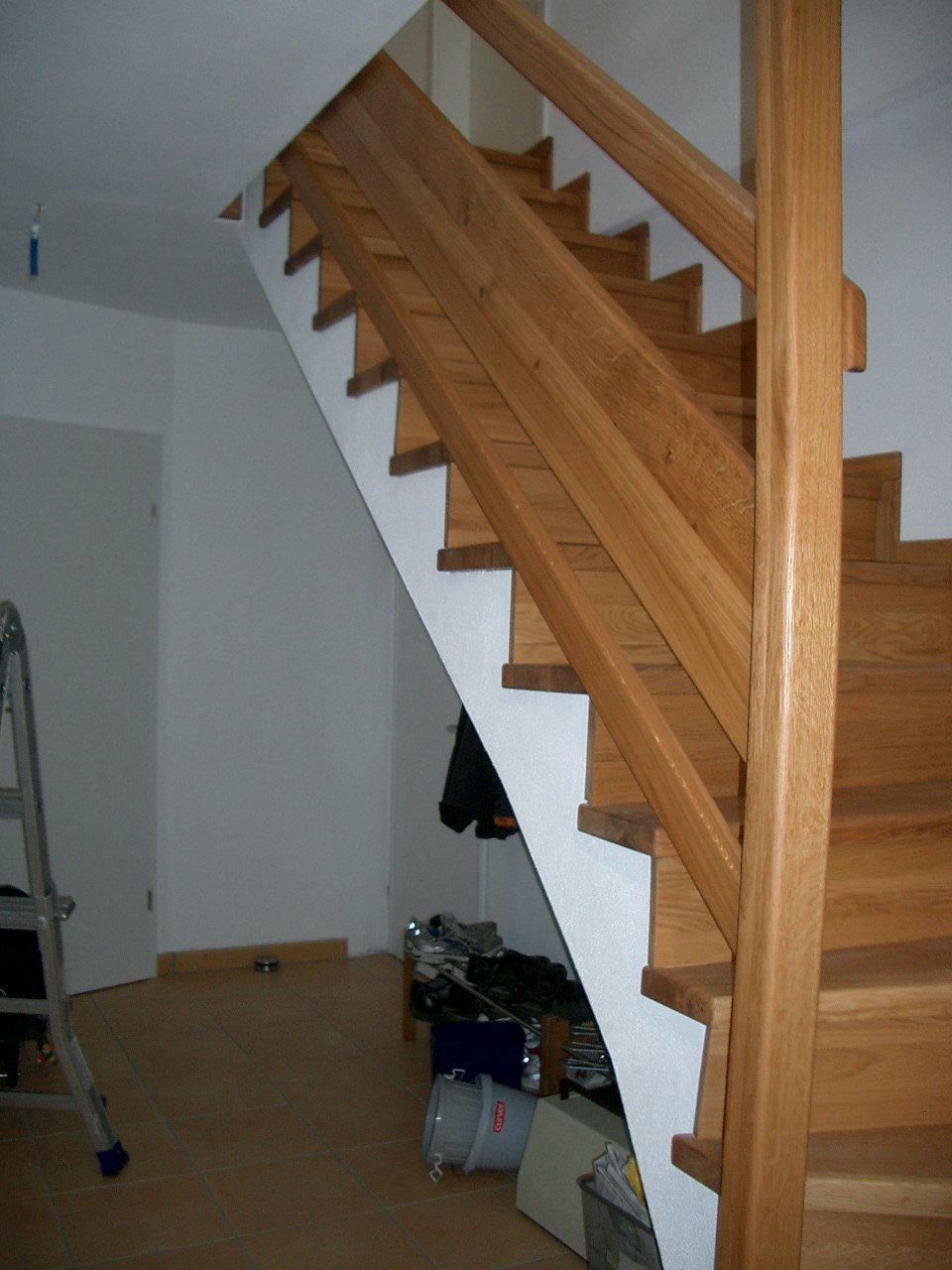 Interior view of a wooden staircase with a white wall and cluttered floor. Items include a folded ladder, a trash bin, boxes, and scattered belongings.