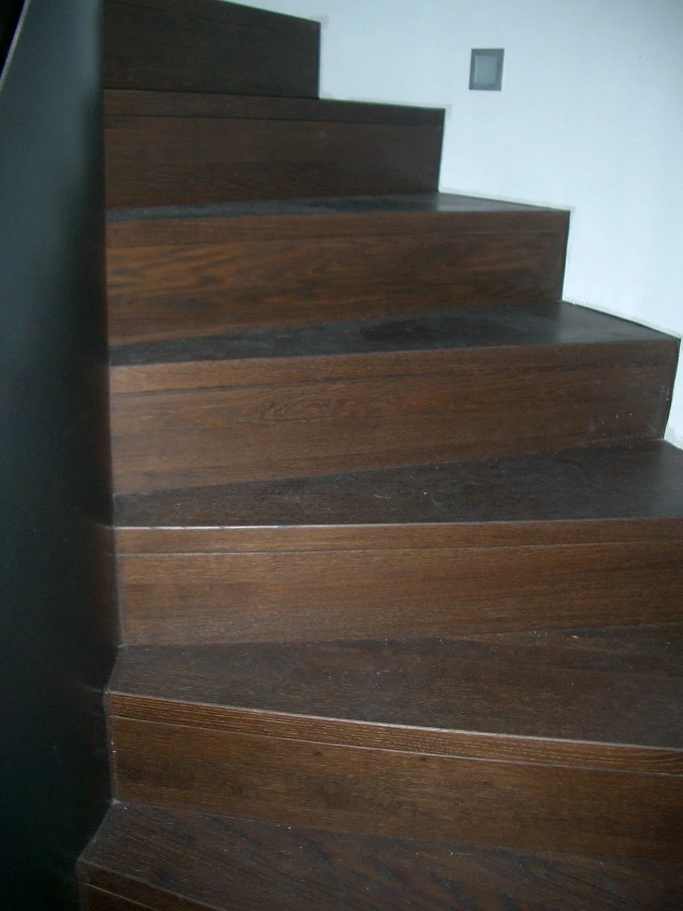 Close-up of wooden staircase with dark brown finish, showing steps ascending to a white wall with a small gray electrical outlet.