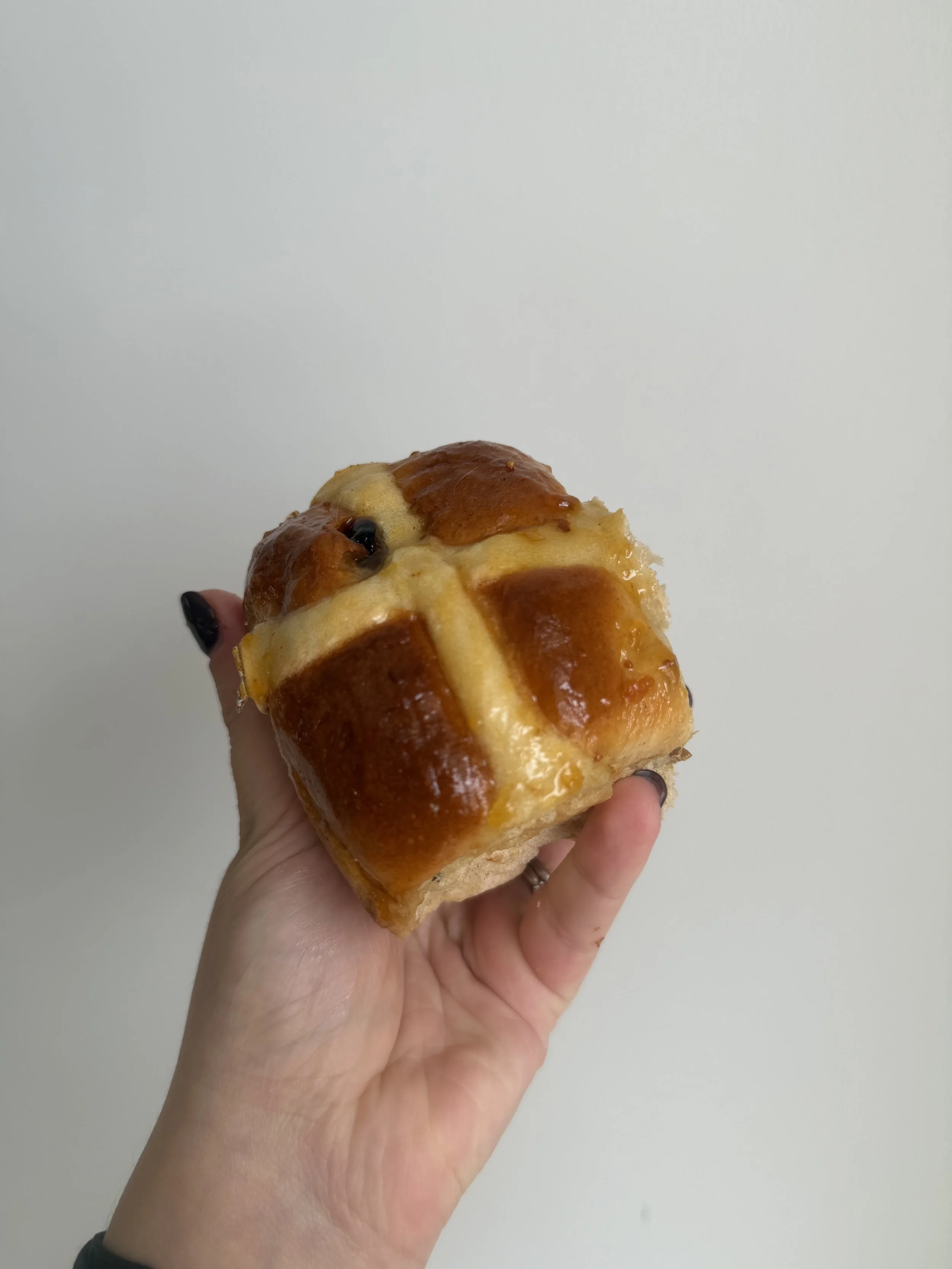 Hand holding a square-shaped bread roll with a cross pattern on top, baked to a golden brown color, against a plain white background.